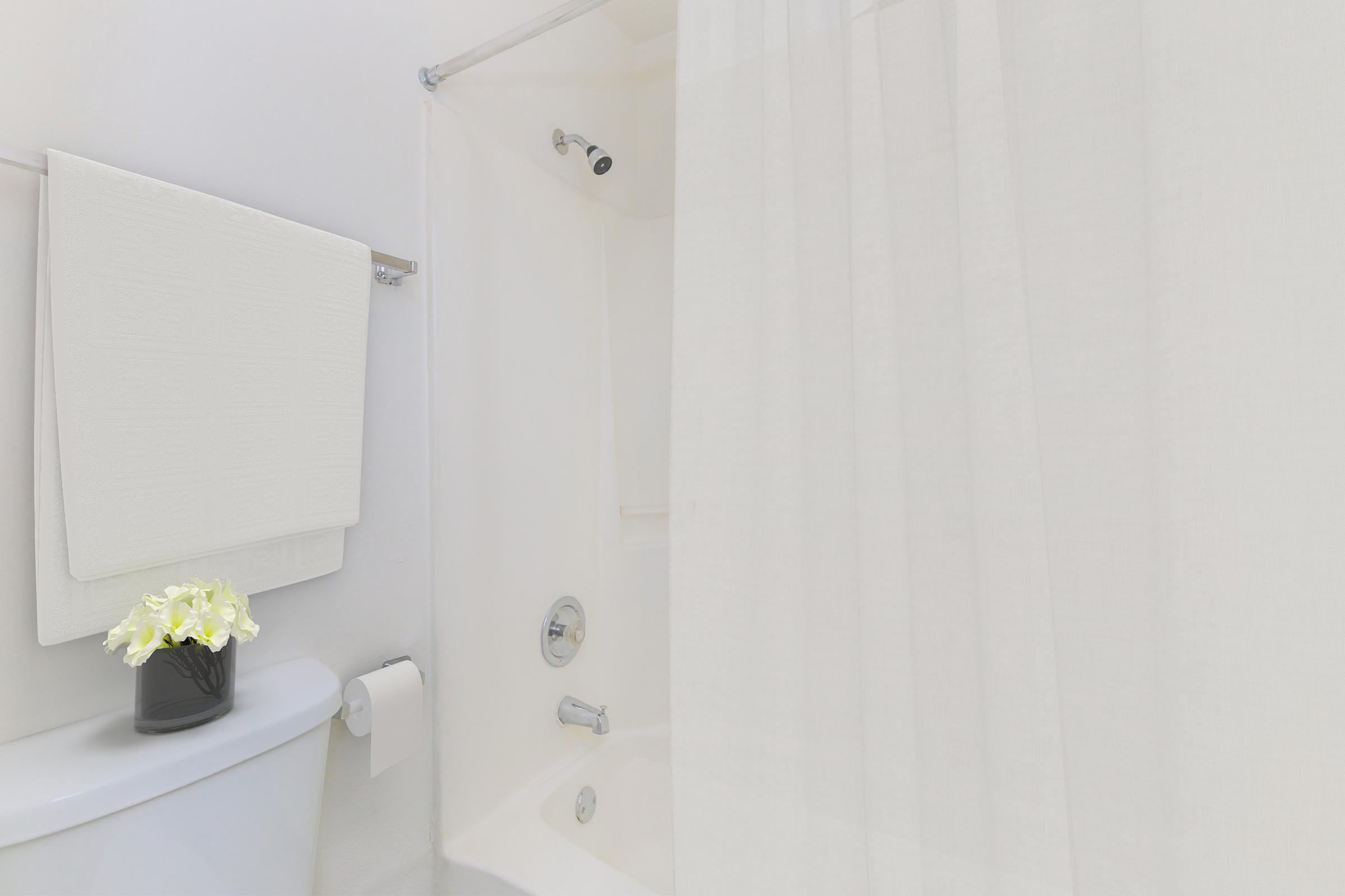 A neatly arranged bathroom featuring a white shower curtain, a towel hanging on a rack, and a potted plant with white flowers on the countertop. The space is clean and minimalistic, creating a soothing atmosphere.