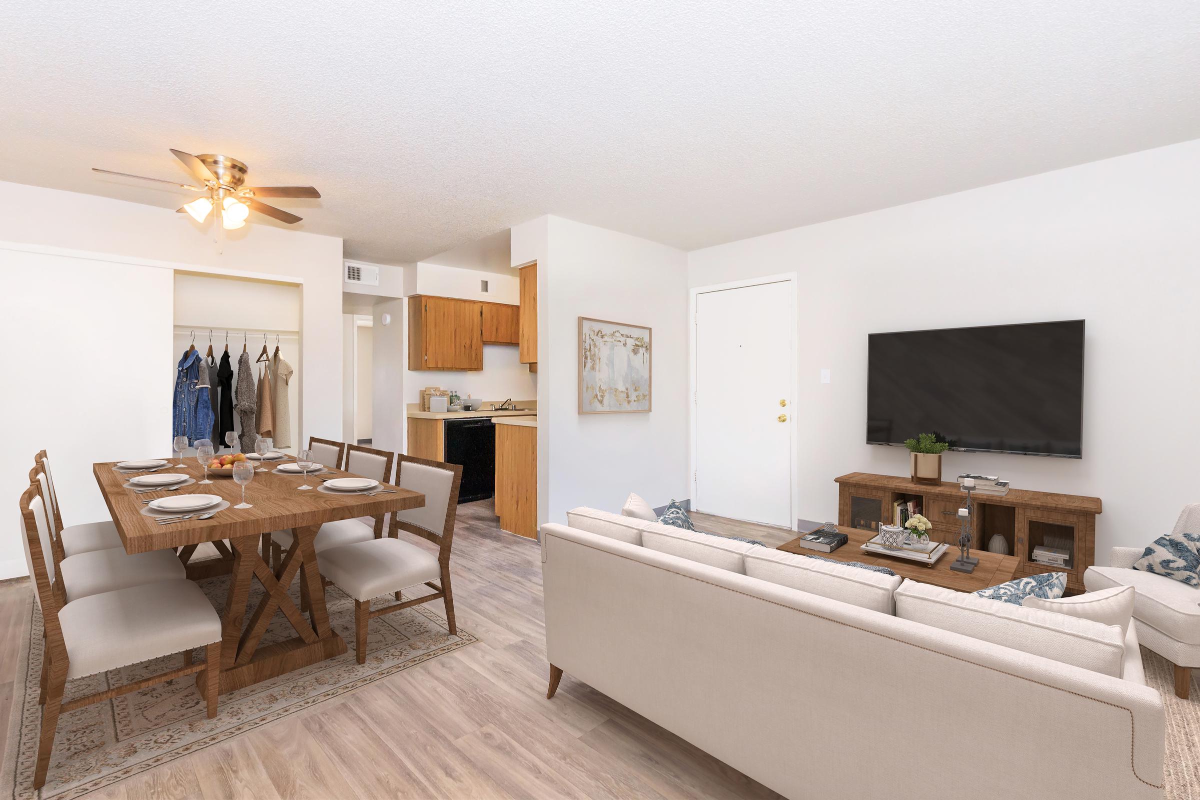A bright and modern living space featuring a dining area with a wooden table set for dinner, comfortable sofa, and a large wall-mounted TV. The kitchen is visible in the background, with wooden cabinets and a doorway leading to a closet with hangers. Natural light fills the room, enhancing the cozy atmosphere.