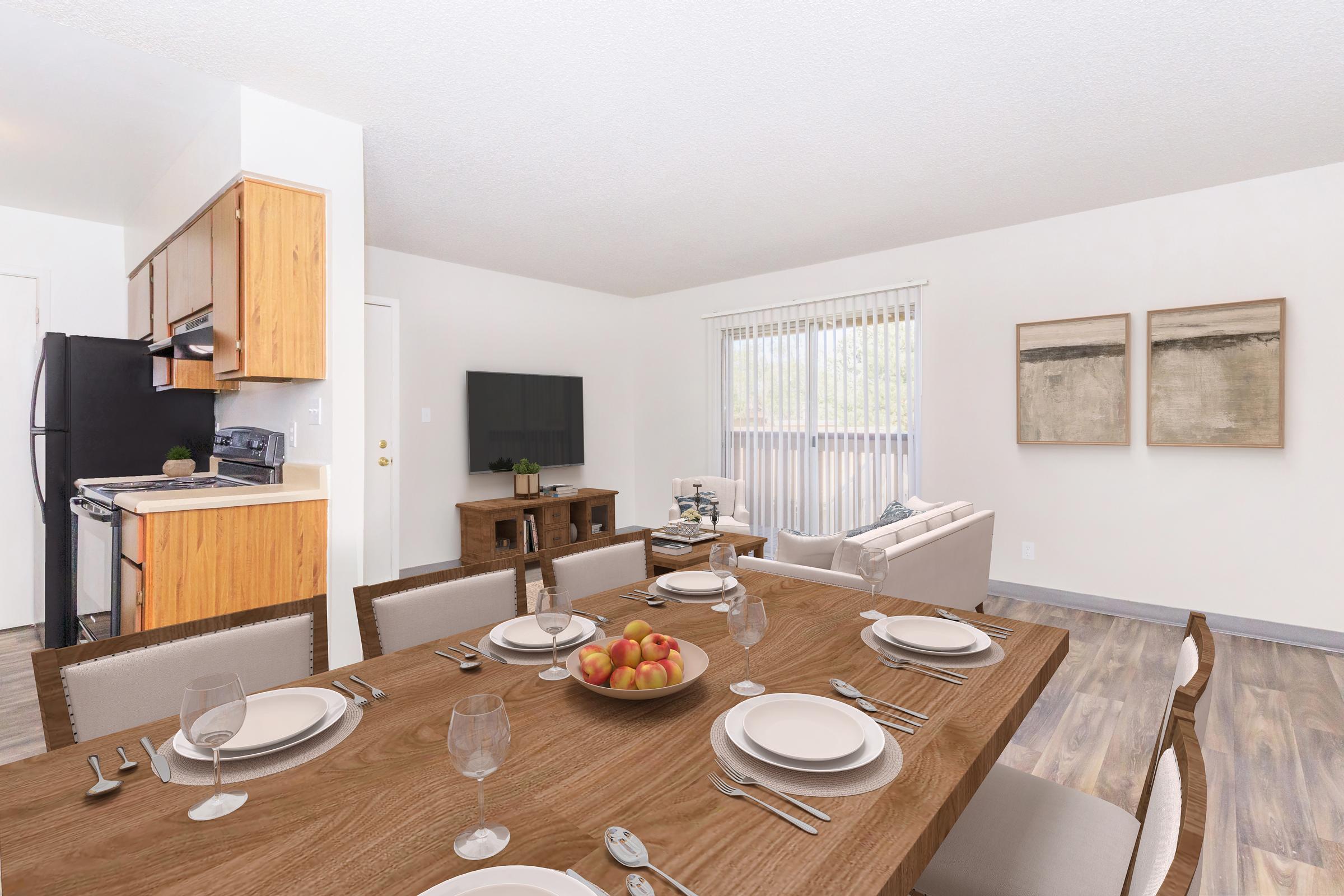 A modern dining area featuring a wooden table set for a meal with plates, glasses, and a bowl of apples. In the background, there is a kitchen with wooden cabinets, a black refrigerator, and a living room area with a TV and a couch. Large windows allow natural light to fill the space.