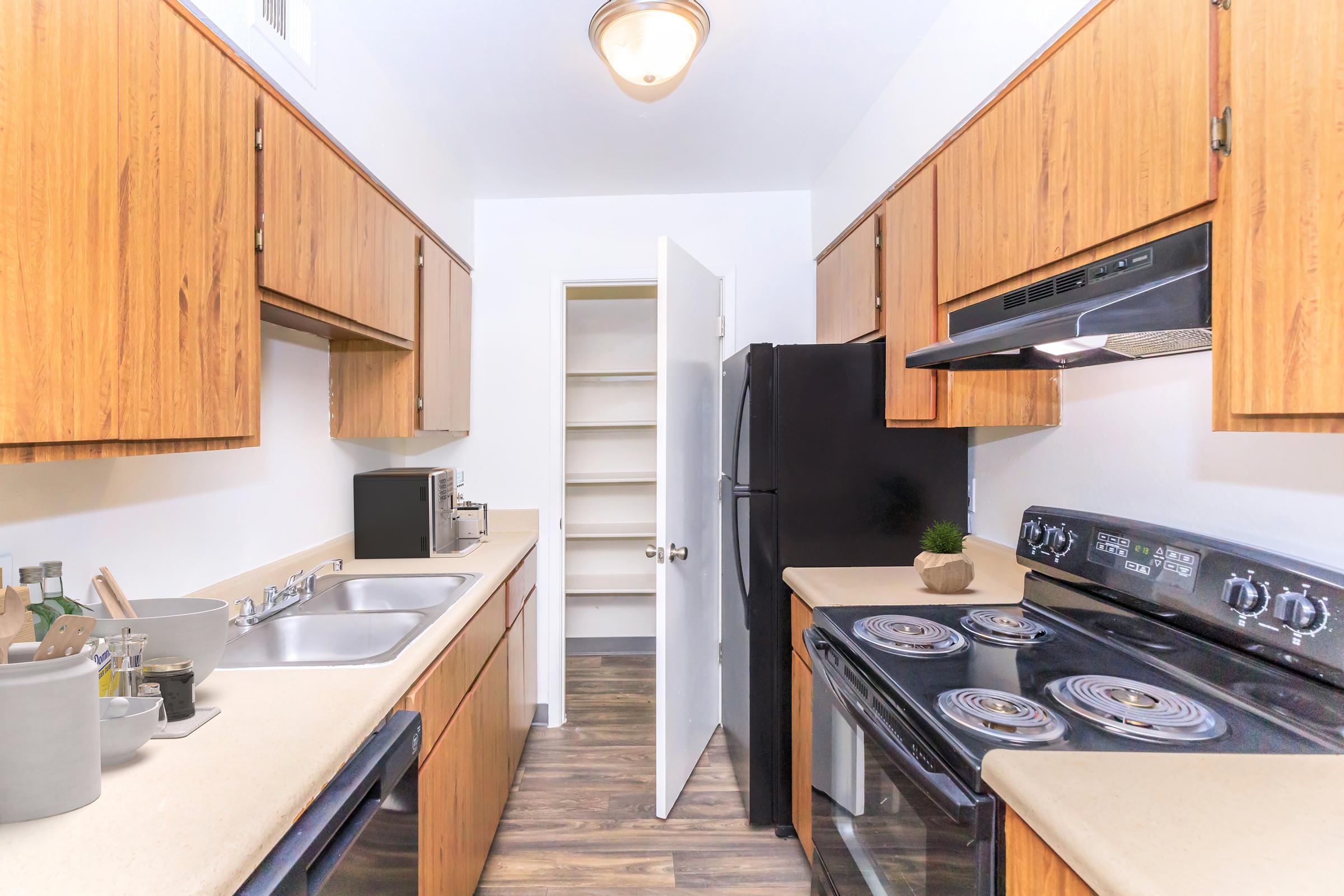 A modern kitchen with wooden cabinets and appliances. The space features a double sink, a stove, and a refrigerator. A door leads to a pantry, and countertop items include kitchen utensils and a small plant. The flooring is a dark wood laminate, giving a contemporary look to the area.