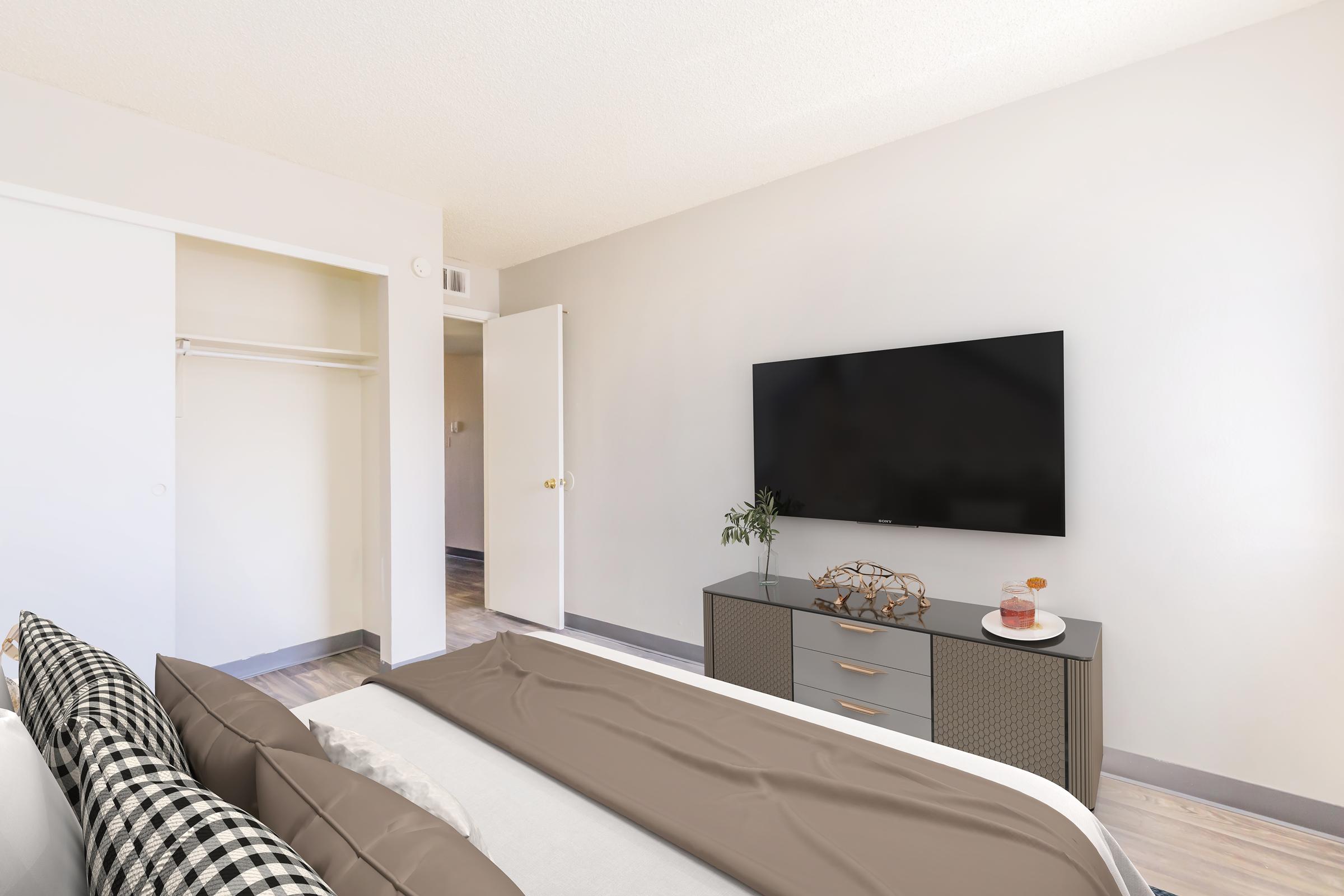 A well-lit bedroom featuring a queen-sized bed with neutral bedding, a sleek gray dresser, and a large flat-screen TV mounted on the wall. A closet is partially visible, and a small plant decorates the dresser, adding a touch of greenery to the modern, minimalist decor.