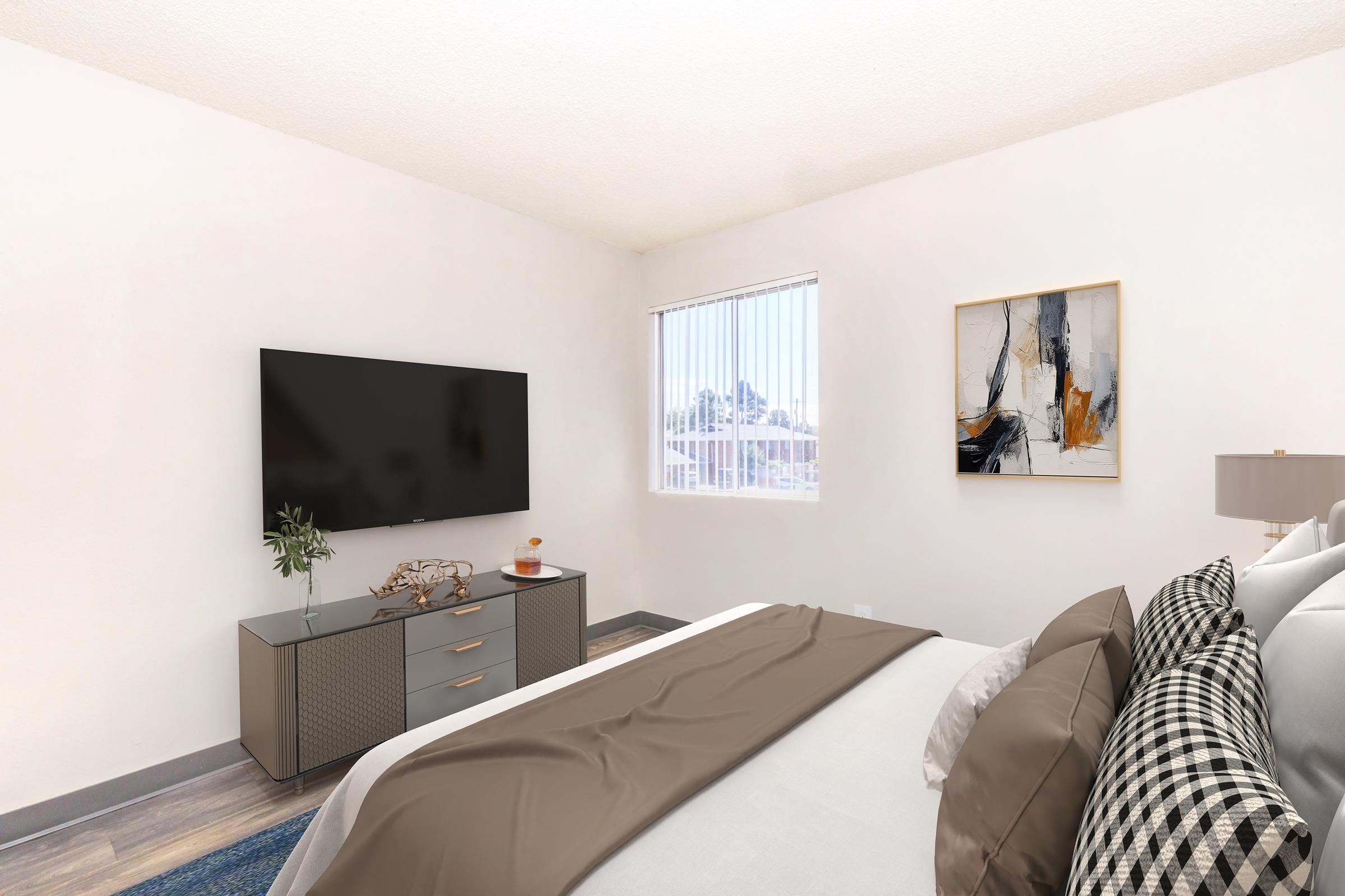 A modern bedroom featuring a neatly made bed with neutral-colored linens, a large flat-screen TV mounted on the wall, a stylish dresser, and an art piece hanging nearby. Large windows with blinds allow natural light to fill the space, creating a bright and inviting atmosphere.