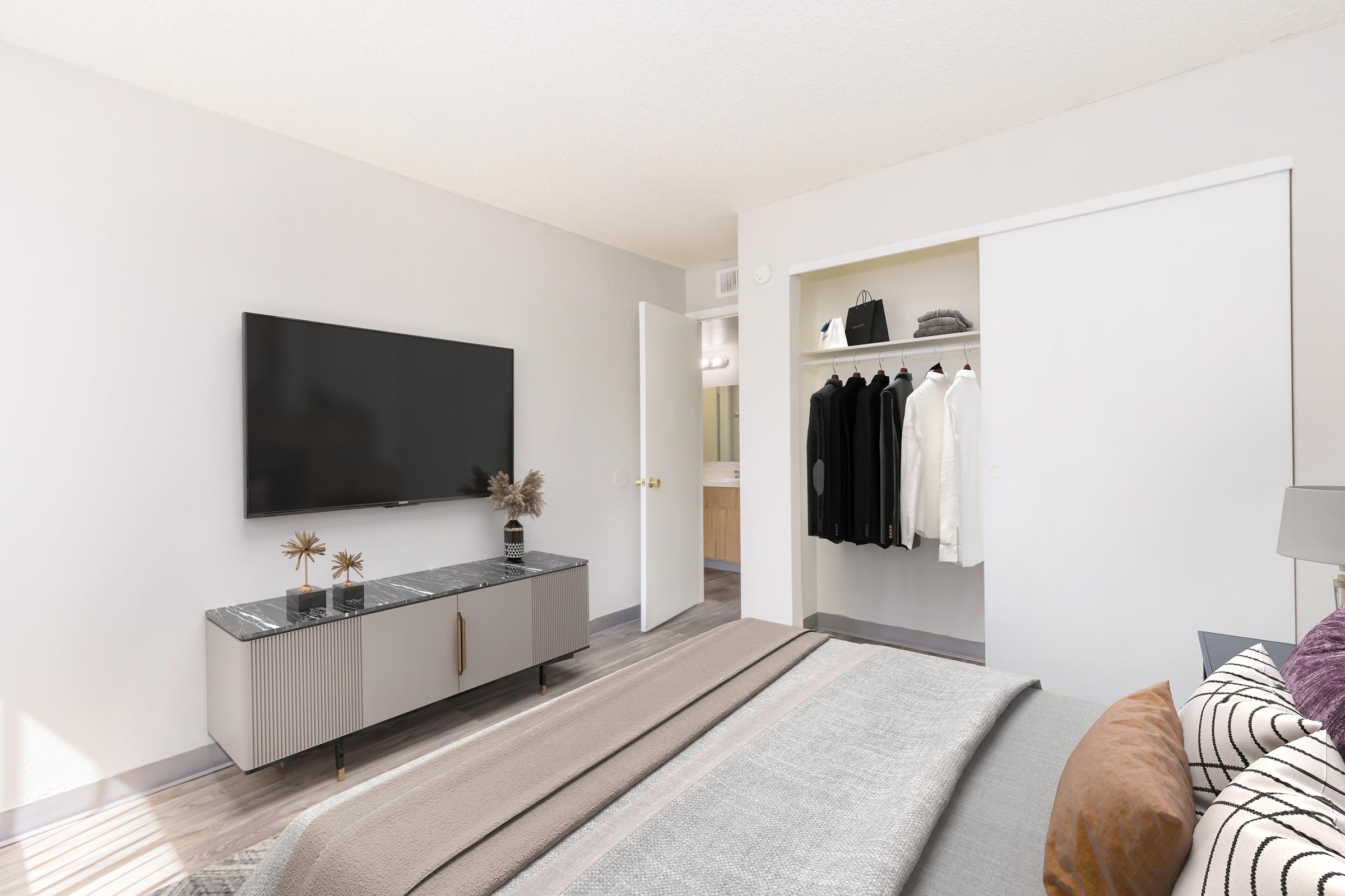 A modern bedroom featuring a bed with a gray blanket, a stylish TV mounted on the wall, and a sleek dresser. A closet with neatly hung clothes is visible through an open sliding door. Soft lighting and minimalistic decor create a cozy atmosphere.