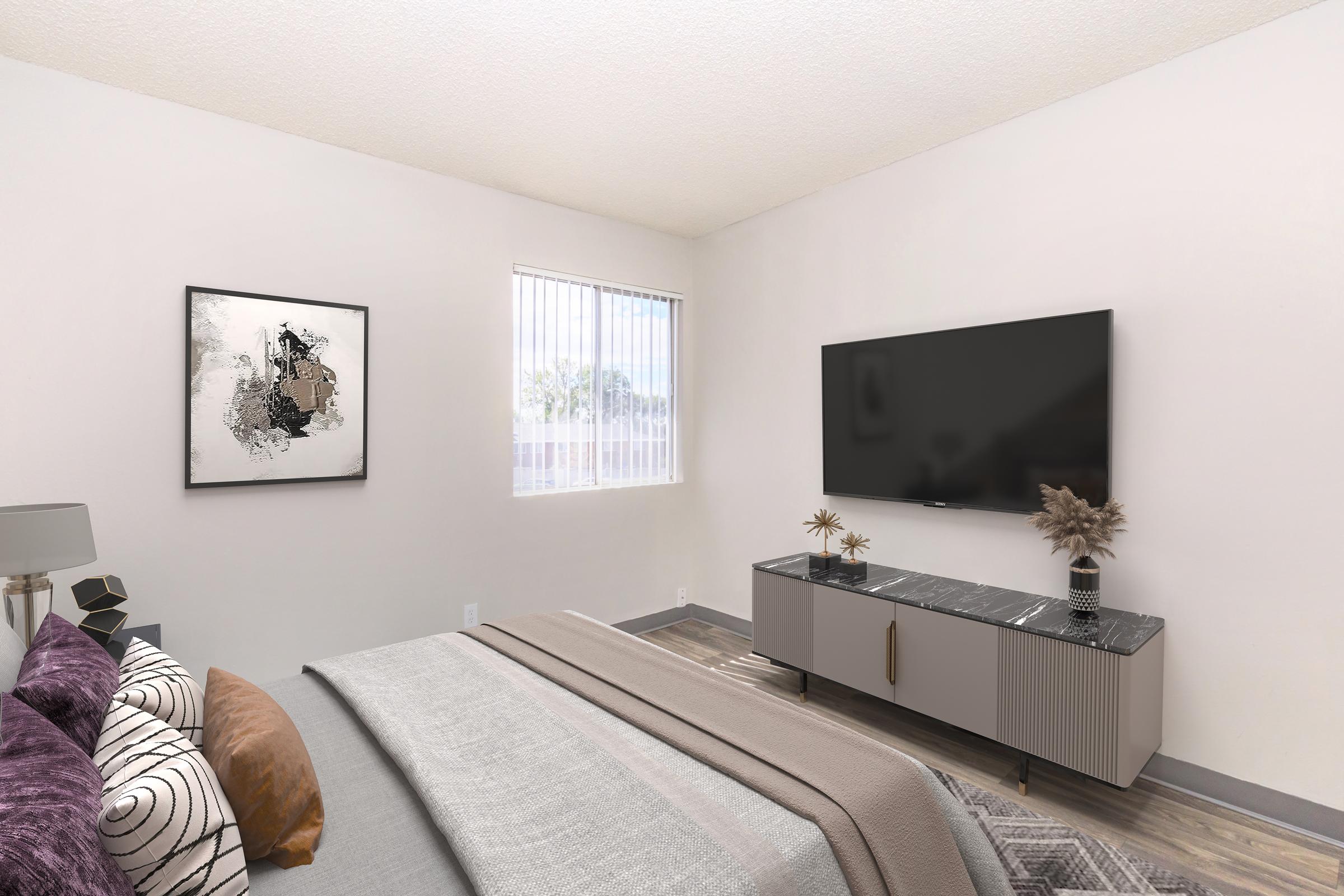 A modern bedroom featuring a neatly made bed with decorative pillows, a stylish gray blanket, a wall-mounted television, and a contemporary TV stand. There's abstract artwork on the wall and a window with vertical blinds, allowing natural light to brighten the space. The flooring is wooden, creating a cozy atmosphere.