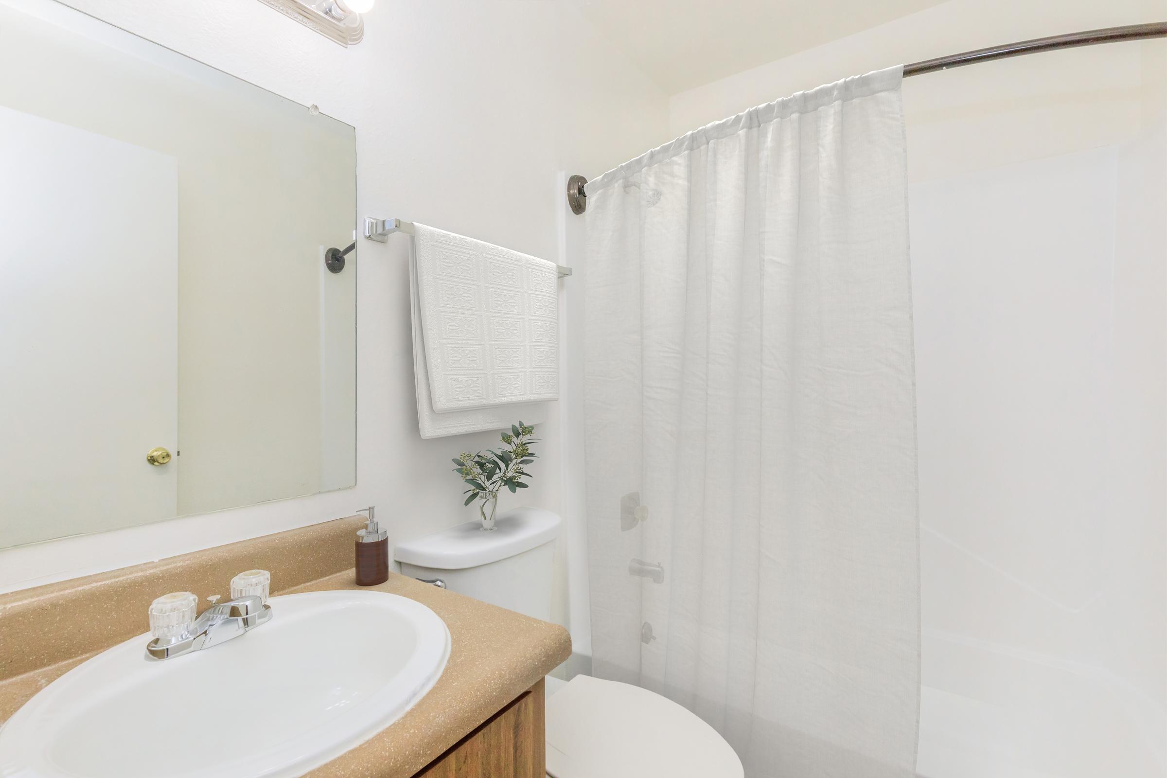 A clean bathroom featuring a round sink with two faucets, a light-colored countertop, a white shower curtain, and a small plant on the counter. A mirror is mounted above the sink, and there's a towel rack with a white towel. The walls are a light color, creating a bright, airy atmosphere.