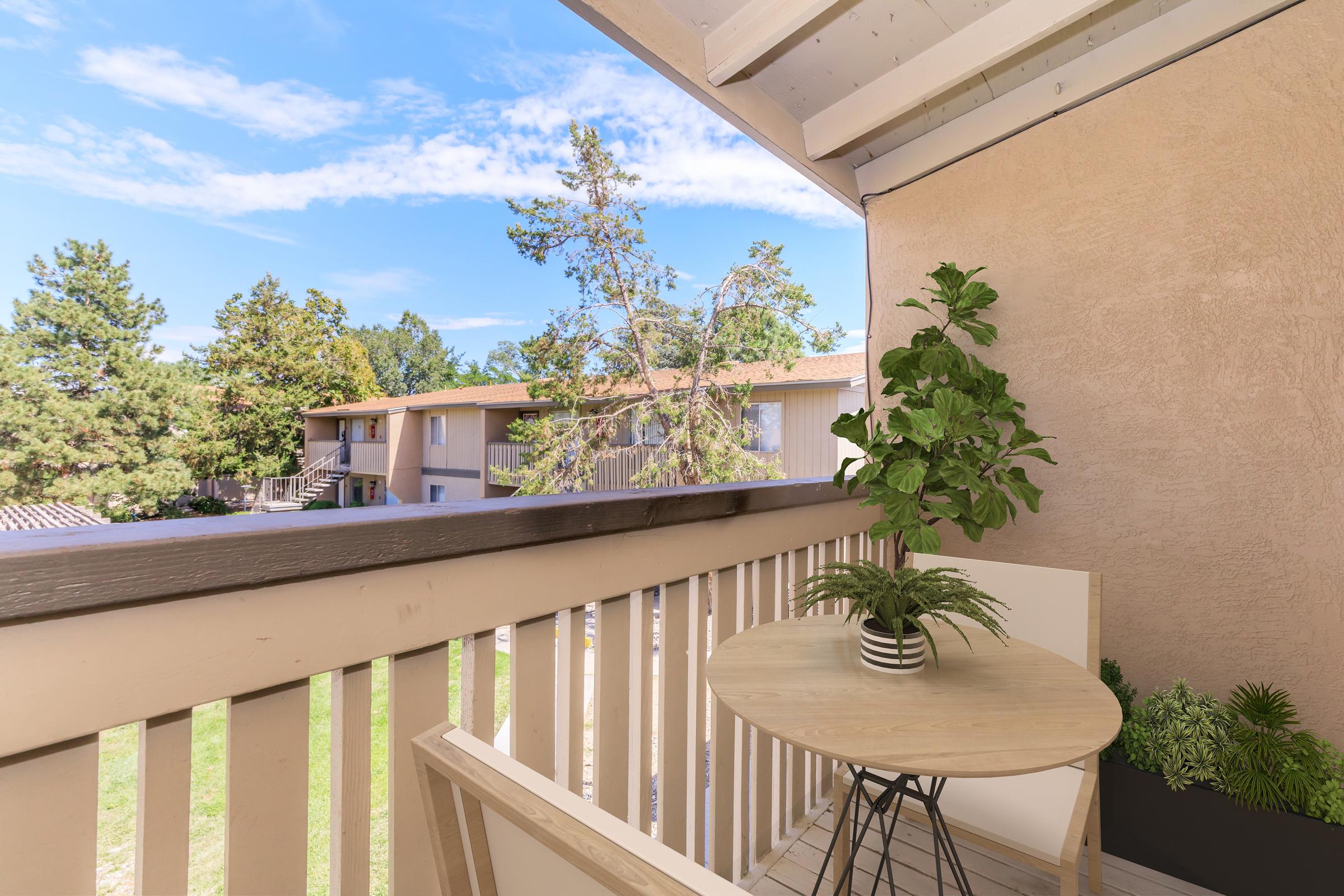 A cozy balcony view featuring a small round table and a potted plant. The railing provides a clear view of a green lawn and nearby apartment buildings surrounded by trees under a bright blue sky.