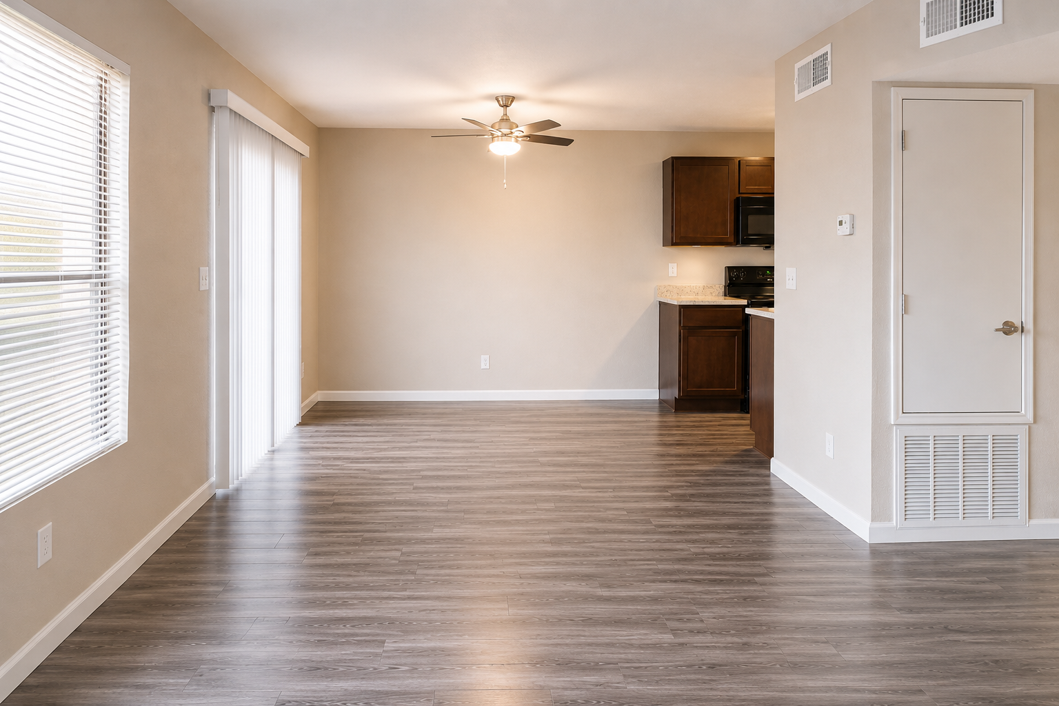Interior view of a modern apartment living space. The room features light-colored walls, a ceiling fan, and a large window with blinds allowing natural light. An open kitchen area with dark wood cabinets and appliances is visible in the background. The flooring is a laminate wood finish, giving a clean and spacious look.