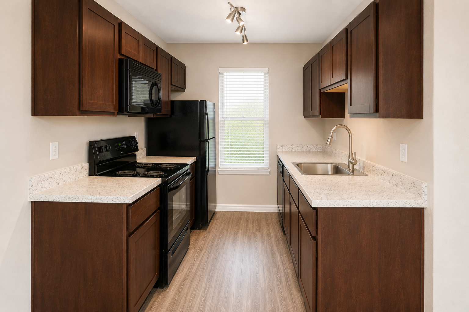 A modern kitchen featuring dark wood cabinets, a black refrigerator, and an oven with a stovetop. The countertops are light-colored, complementing the cabinetry. A window allows natural light, and there's a stainless steel sink with a faucet. The flooring is a warm wood-like laminate.