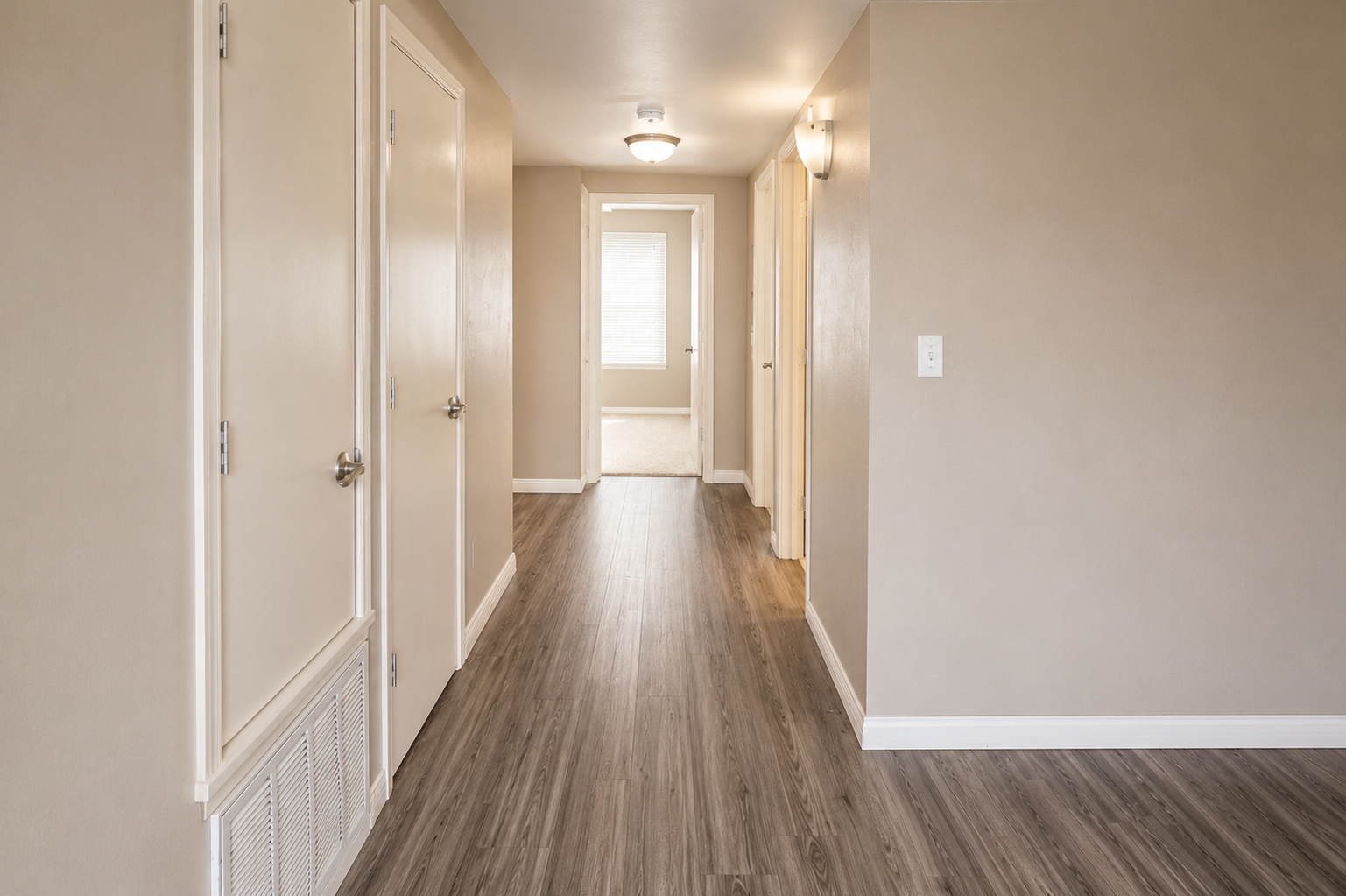 A well-lit hallway featuring beige walls and wooden flooring. The hallway has several closed doors on either side, leading to different rooms, and ends with a door that opens to a bright light. A ceiling light fixture illuminates the space, enhancing the modern and inviting atmosphere.