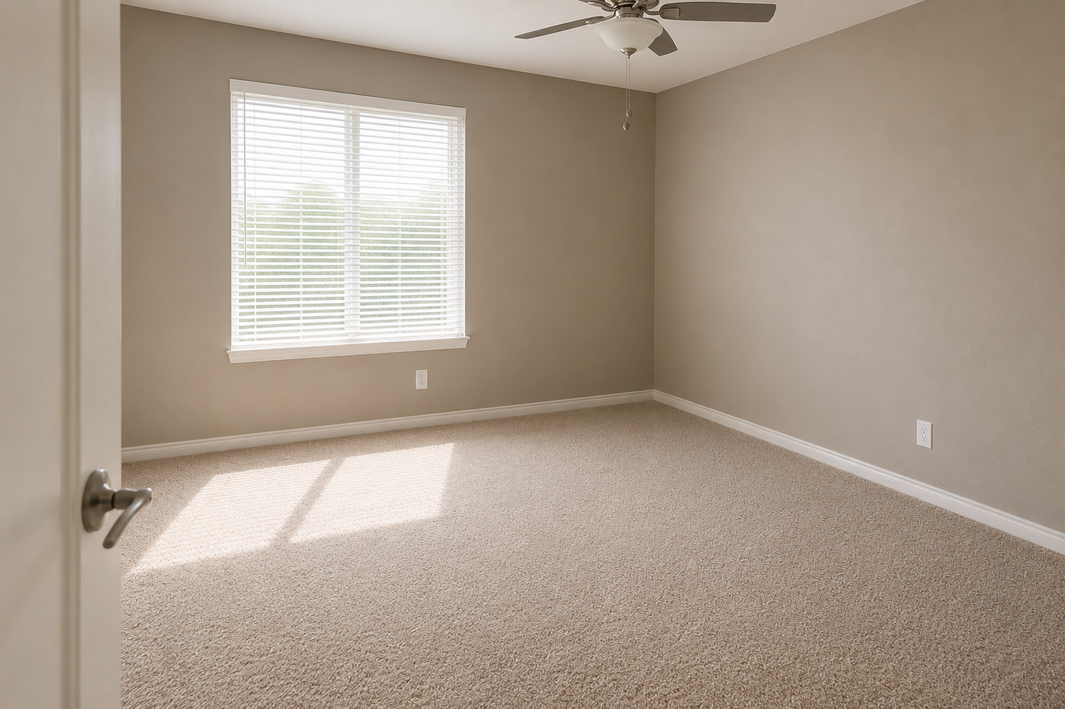 A vacant room with beige walls and carpet. There is a window with white blinds allowing natural light to enter, casting a shadow on the floor. A ceiling fan is mounted above. The room appears clean and uncluttered, suitable for various uses.