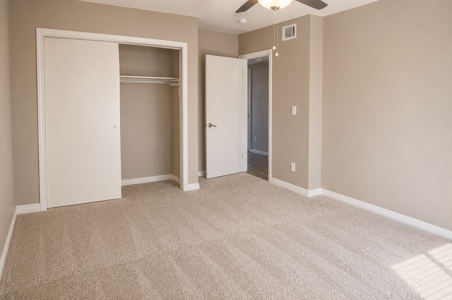 A spacious room with light beige walls and plush carpet. It features a closet with sliding doors, an open doorway leading to another space, and a ceiling fan. The room is well-lit by natural light, creating a bright and inviting atmosphere.