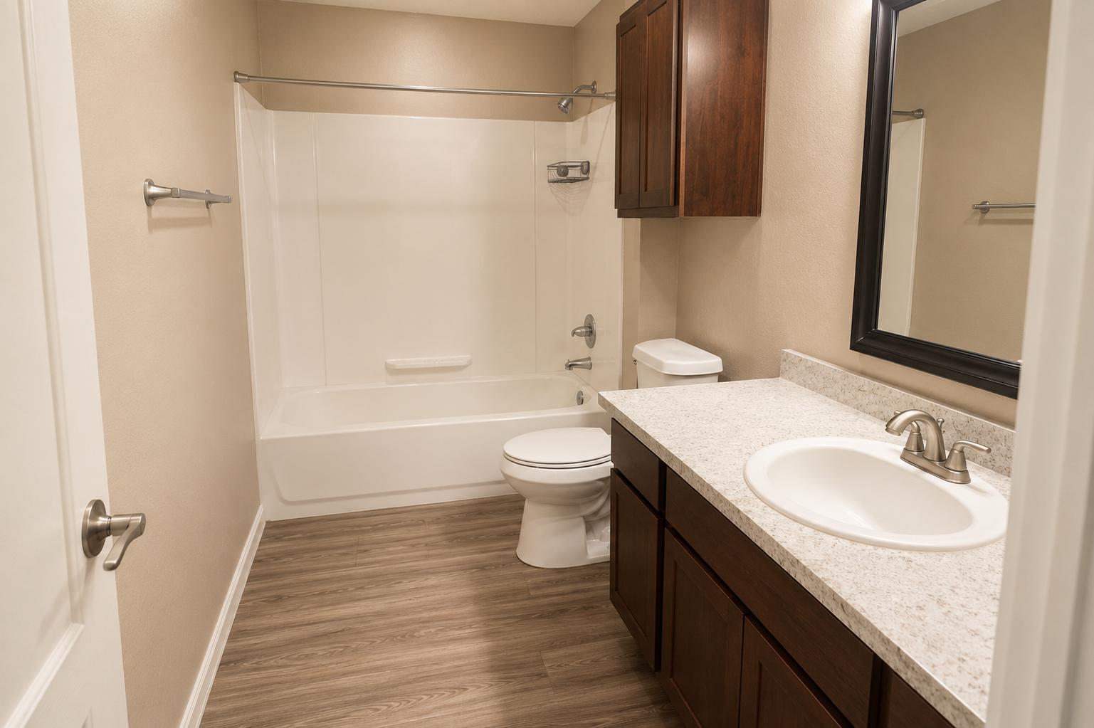 A clean and modern bathroom featuring a bathtub with a shower, a toilet, and a sink with a countertop. The walls are painted light, and there are wooden cabinets for storage. A large mirror hangs above the sink, and the floor is finished with wood-like vinyl.