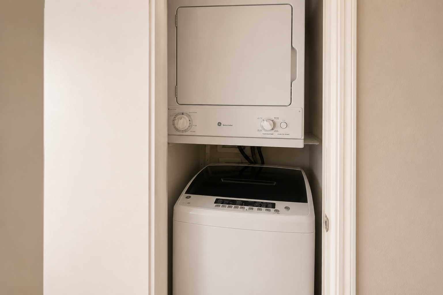 A compact laundry area featuring a top-loading washing machine and a stacked dryer inside a closet. The appliances are white with control panels visible, showcasing their modern design and space-saving arrangement. The background is a soft beige color, giving a warm and neutral tone to the space.