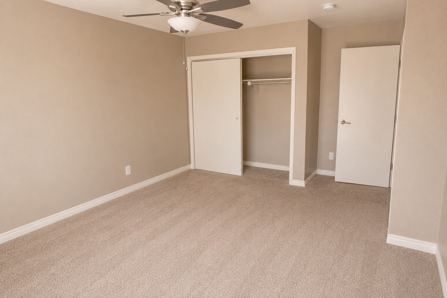 A neutral-toned empty bedroom with beige walls and carpet. The room features a ceiling fan, a built-in closet with an open sliding door, and a door leading to another area. Natural light enters through the doorway, creating a spacious and clean atmosphere.