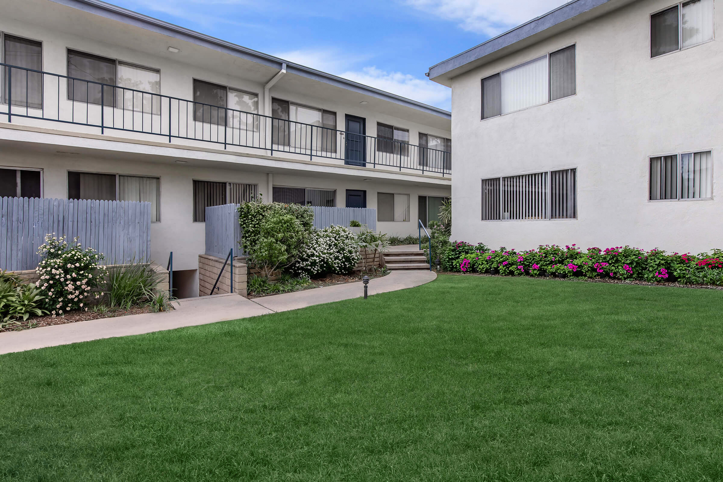 A view of a residential building complex featuring two multi-story buildings with balconies, surrounded by neatly maintained gardens and flower beds. A green lawn is in the foreground, and a path leads to a staircase. The sky is clear with scattered clouds.