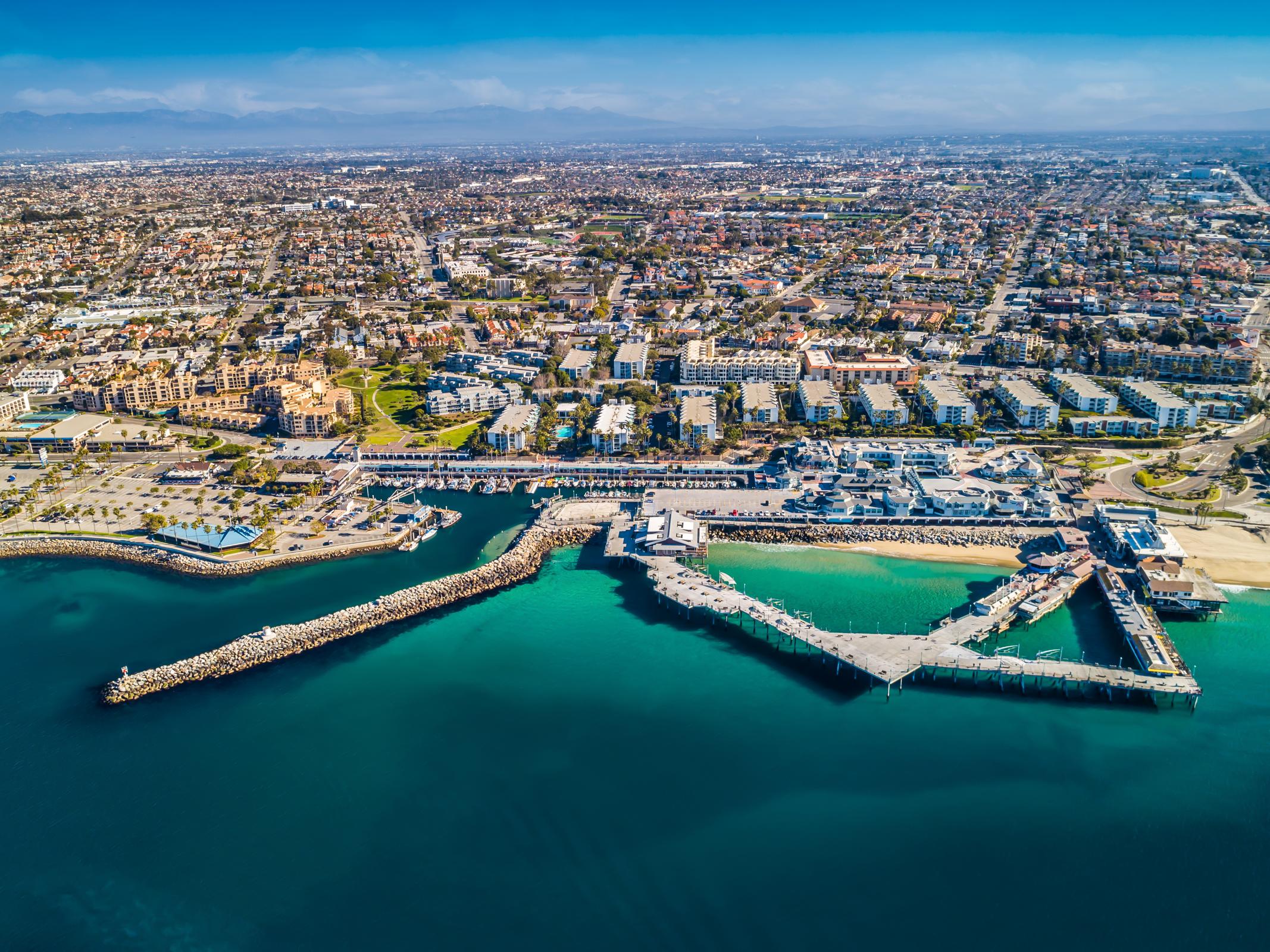 Aerial view of a coastal city, showcasing a marina with several piers extending into turquoise waters. The shoreline features sandy beaches and a mix of residential and commercial buildings. In the background, a landscape of greenery and urban development is visible under a clear blue sky.