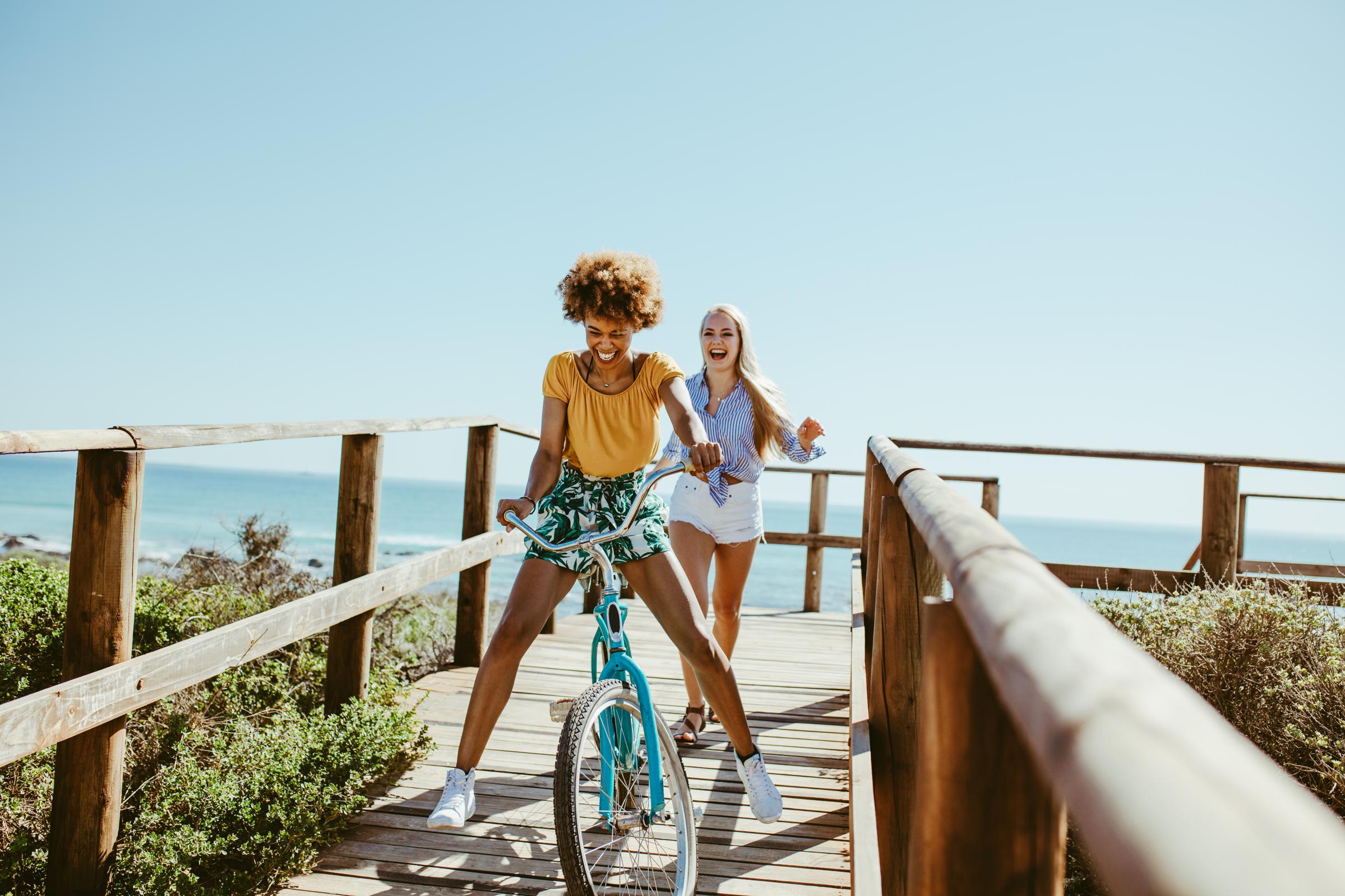 Two women enjoying a sunny day by the beach. One woman with curly hair rides a blue bicycle on a wooden pathway, while the other woman, blonde and smiling, runs beside her. The background features a clear blue sky and calm ocean waves.