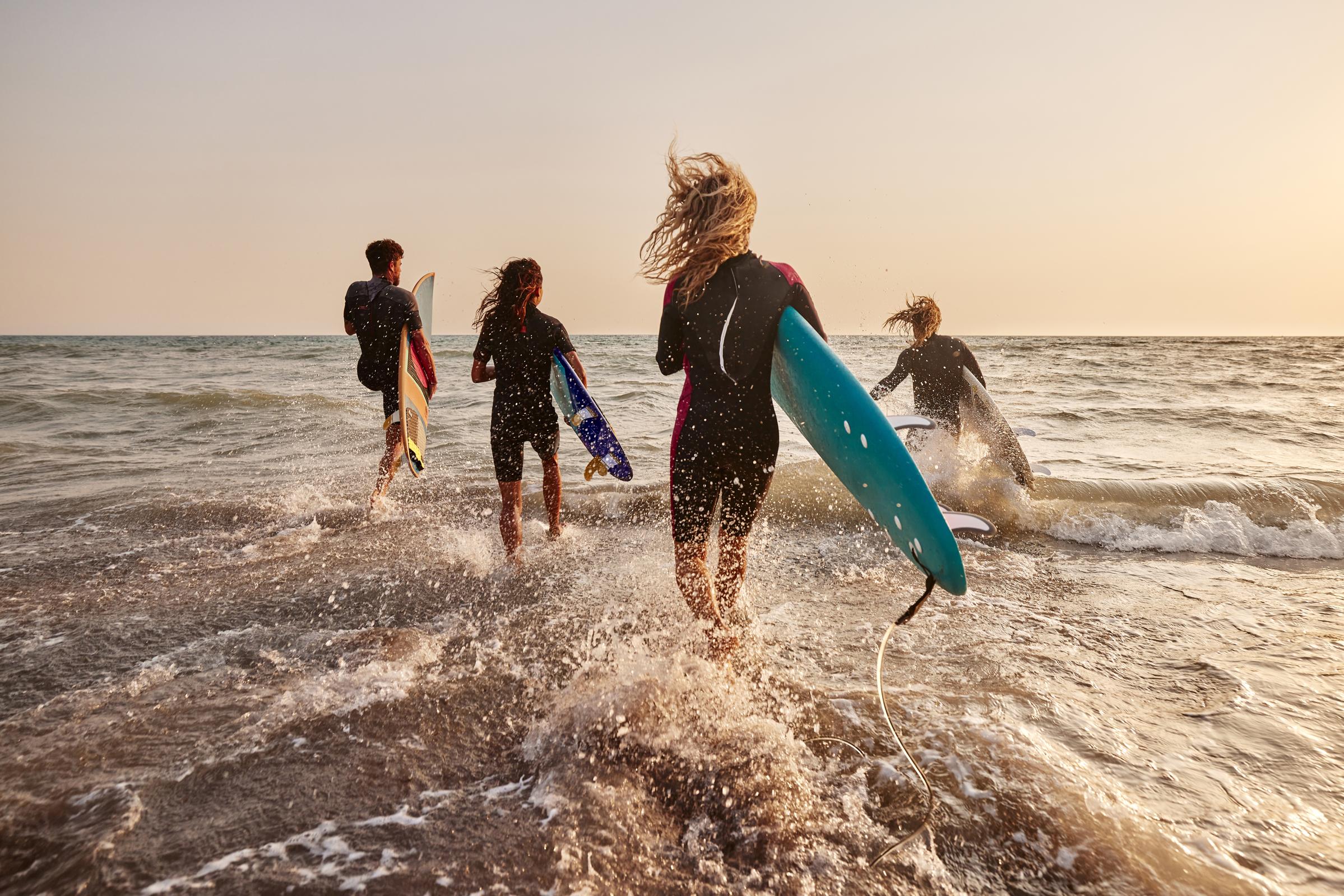 A group of four surfers, two women and two men, running toward the ocean with surfboards in hand. The background features gentle waves and a sunset, casting warm hues over the scene. The surfers are dressed in wetsuits, and the atmosphere is lively and energetic, capturing the excitement of a beach day.
