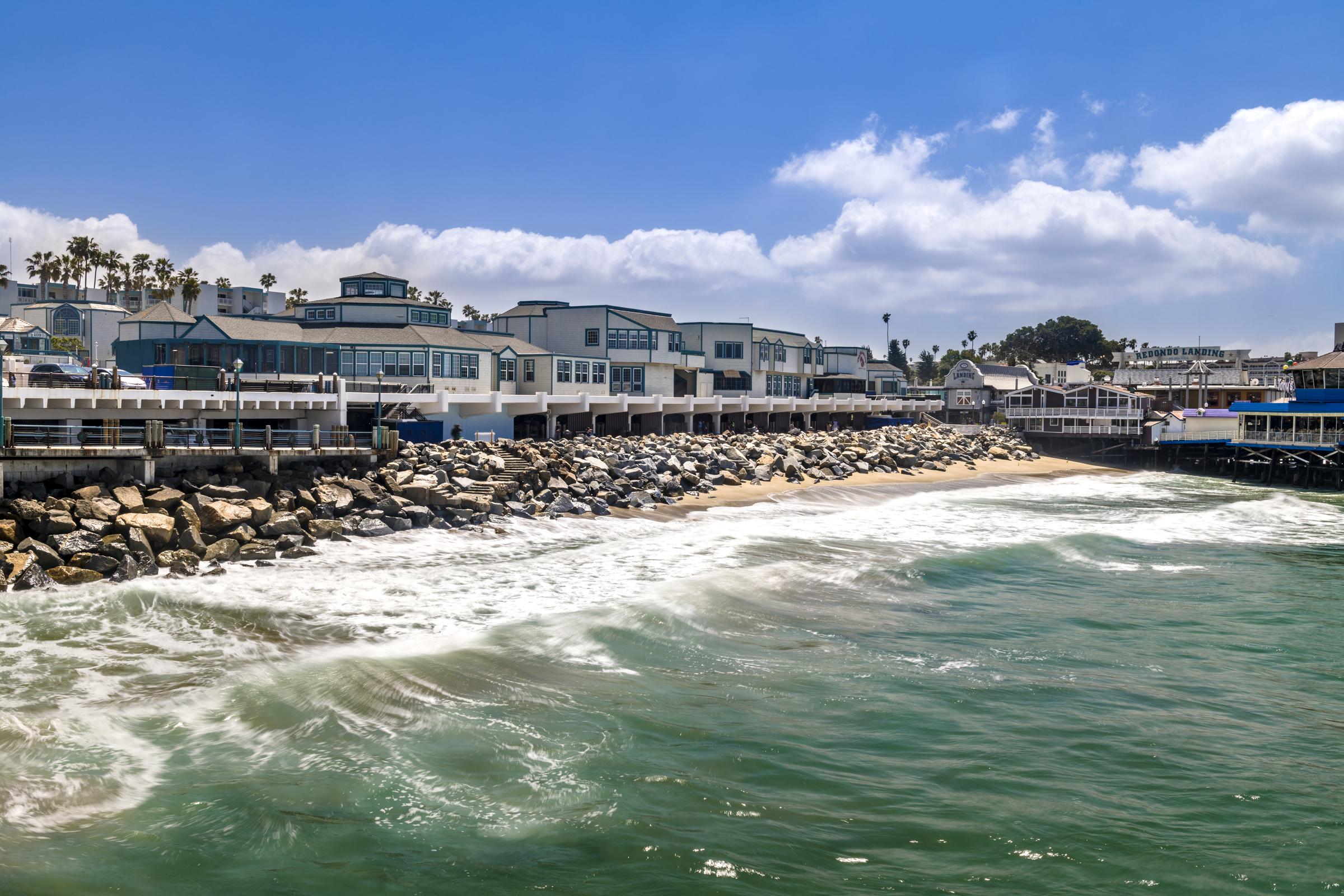Coastal view featuring a shoreline with gentle waves lapping against rocky outcroppings. In the background, modern buildings line the waterfront under a bright blue sky with scattered clouds. The scene captures a serene beach atmosphere, ideal for leisurely strolls and enjoying the seaside.