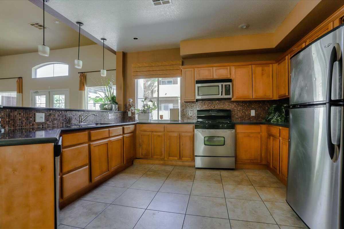 Spacious kitchen featuring wooden cabinets, a stainless steel refrigerator, and an oven. The countertops are dark, complemented by stylish tile backsplash. Natural light floods the room through large windows, enhancing the inviting atmosphere. Modern light fixtures hang above the island area.