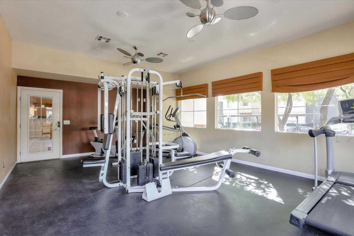 A fitness room featuring various exercise equipment, including a treadmill, elliptical machine, and multi-station weight system. The space has large windows with shades and ceiling fans, creating a bright and airy atmosphere suitable for workouts.