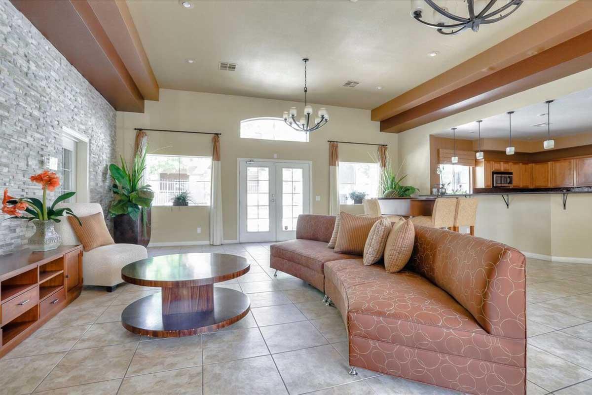 A modern living area featuring a curved brown sofa, a round coffee table, and decorative plants. The walls display a stone texture, and large windows allow natural light. A dining area with bar stools is visible in the background, along with pendant lighting and warm color accents.