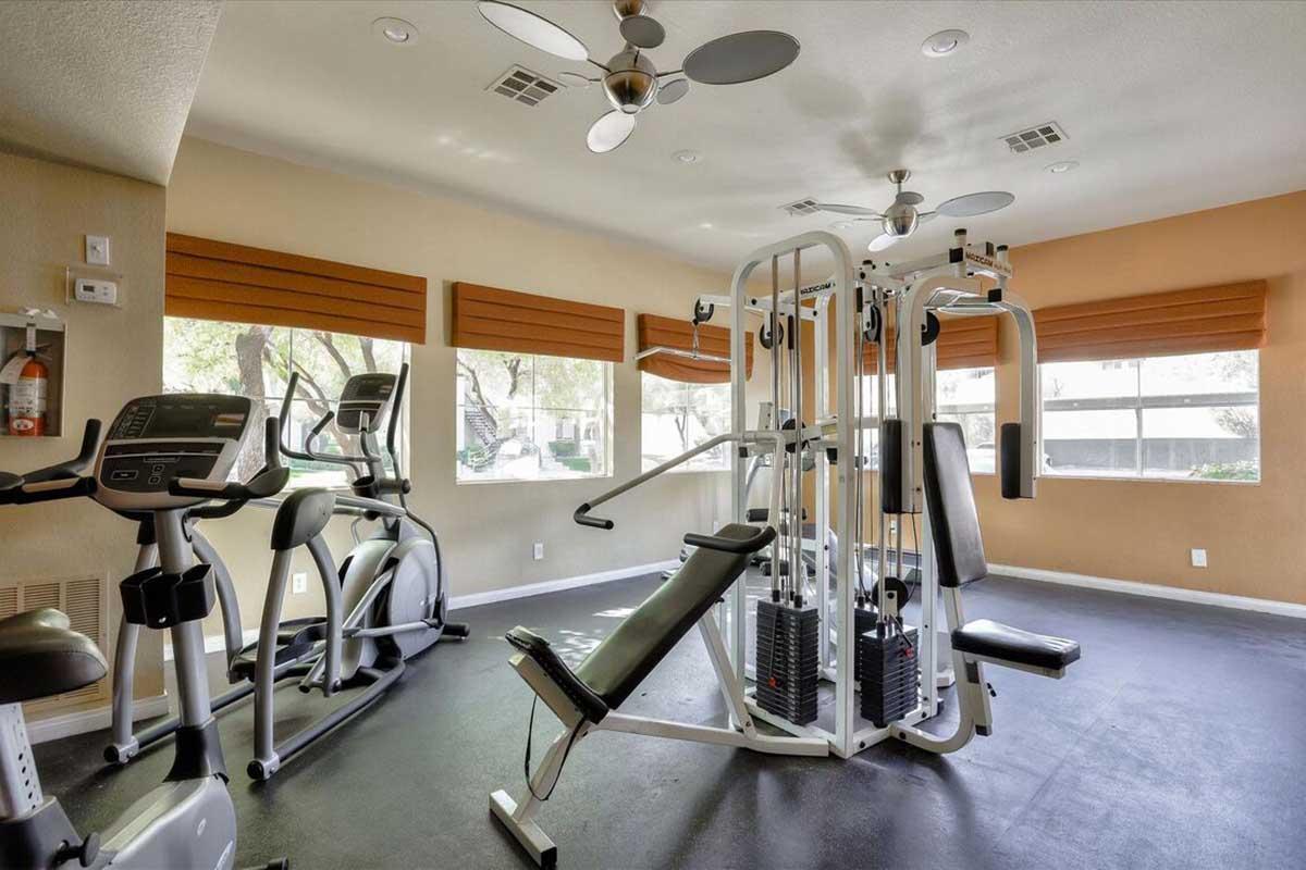 Interior view of a small gym featuring various fitness equipment, including cardio machines like treadmills and ellipticals, along with a multi-station weight machine. The room has large windows with shades, allowing natural light, and has a clean, spacious layout. Ceiling fans are visible.