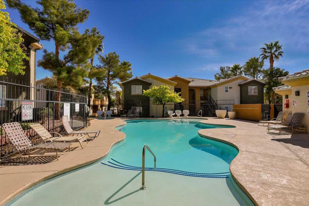 A serene swimming pool area surrounded by lush palm trees and colorful apartment buildings. The pool features a curvy design with a diving ladder, lounge chairs positioned nearby, and clear blue skies in the background, creating a tranquil and inviting atmosphere for relaxation.