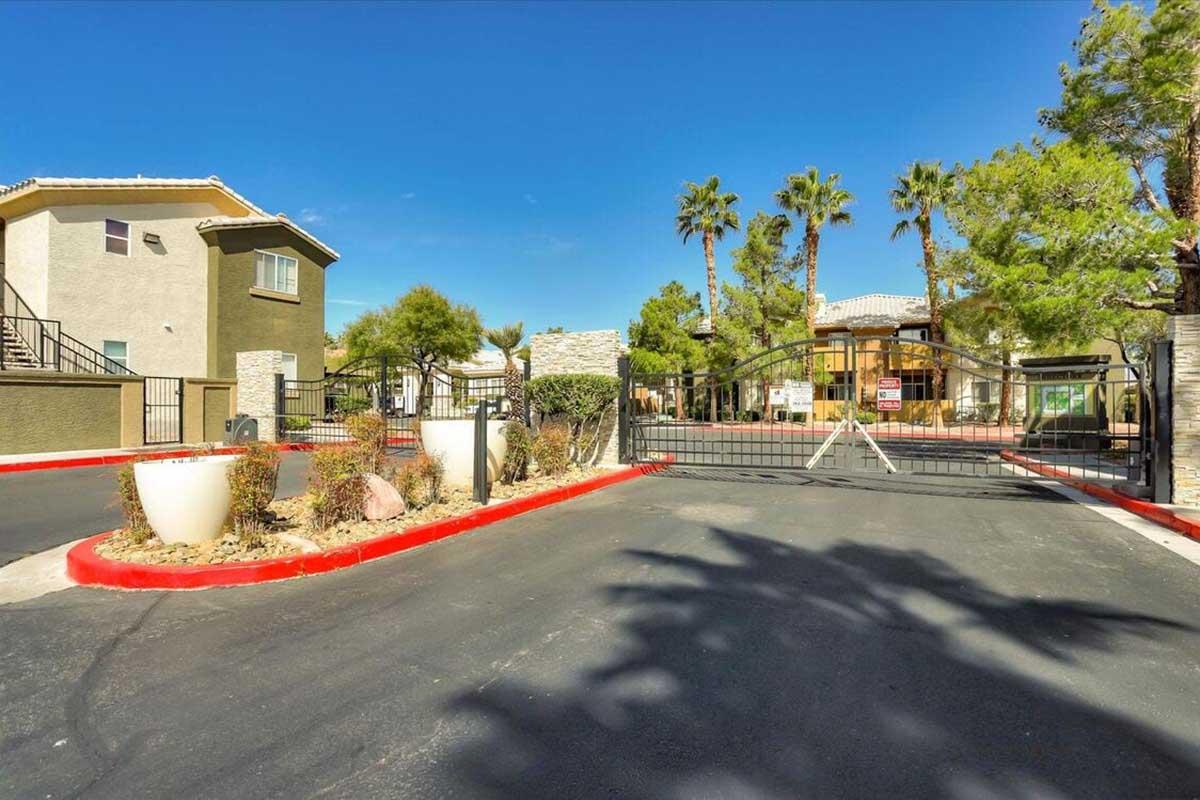 A gated entrance to a residential complex featuring a paved driveway lined with palm trees and landscaped planters. Buildings in the background include two-story structures with balconies, under a clear blue sky. The entrance is marked with a security gate, enhancing privacy and security.