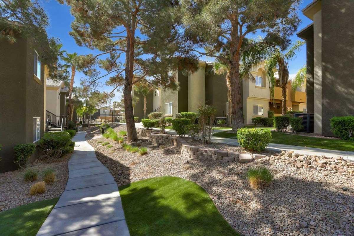 A landscaped pathway meanders through a residential area, flanked by green lawns and bushes. Palm trees and other foliage provide shade, while buildings with light-colored facades stand in the background, creating a tranquil and inviting atmosphere.