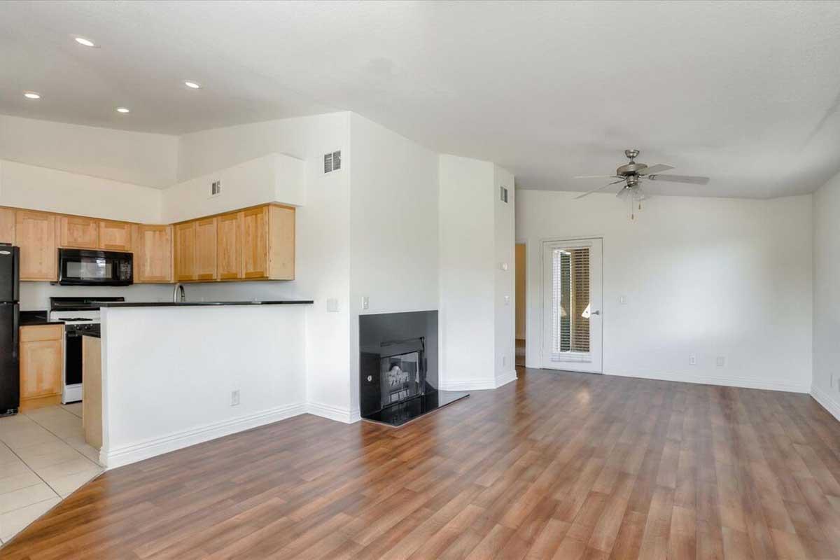 Spacious living room with hardwood floors, a ceiling fan, and a fireplace. The adjacent kitchen features wooden cabinets and modern appliances. Natural light enters through the sliding doors leading to the outside, creating a bright and inviting atmosphere.