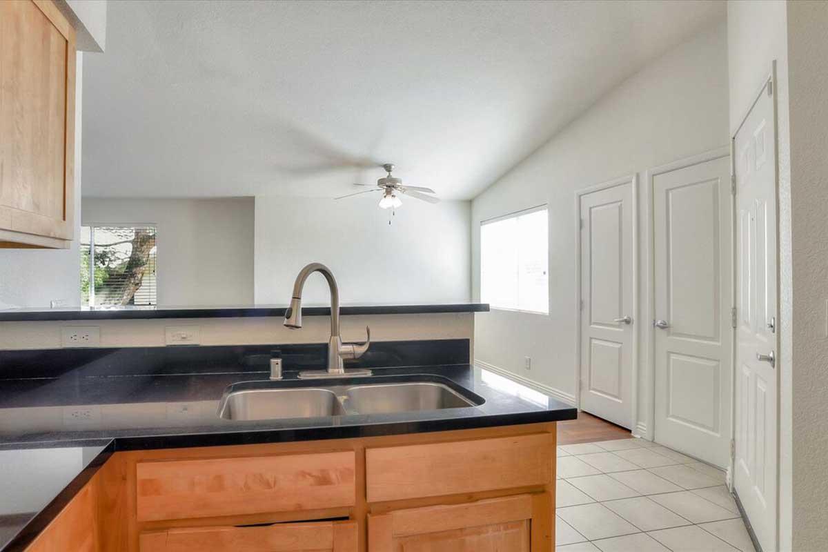 View of a modern kitchen featuring a black countertop, wooden cabinets, and a stainless steel sink. A ceiling fan is visible, along with a window allowing natural light. The background shows an adjoining living space with white walls and doors.