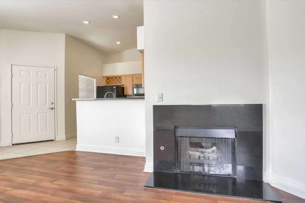 Interior view of a cozy living space featuring a modern fireplace with a black surround, light hardwood flooring, and a kitchen area visible in the background. A white door leads outside, and natural light enters through a window, creating a warm atmosphere.