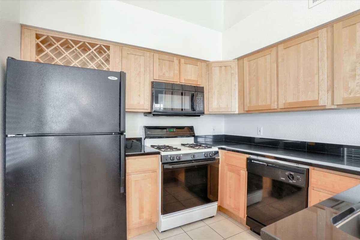 Modern kitchen featuring wooden cabinetry, a black refrigerator, a built-in microwave, and a gas stove with a stainless steel oven. The counters are dark granite, and the flooring is tiled. A wine rack is visible above the cabinets, providing additional storage space.