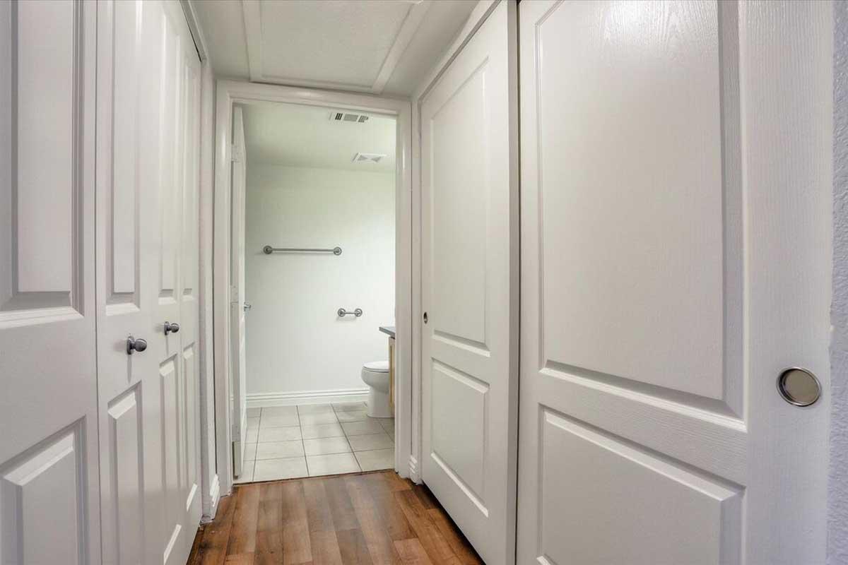 A narrow hallway with white walls and doors on either side leading to a bathroom. The floor is wooden, and tiles are visible at the end of the hallway. A toilet and grab bar can be seen in the bathroom area. The space is well-lit and modern in design.