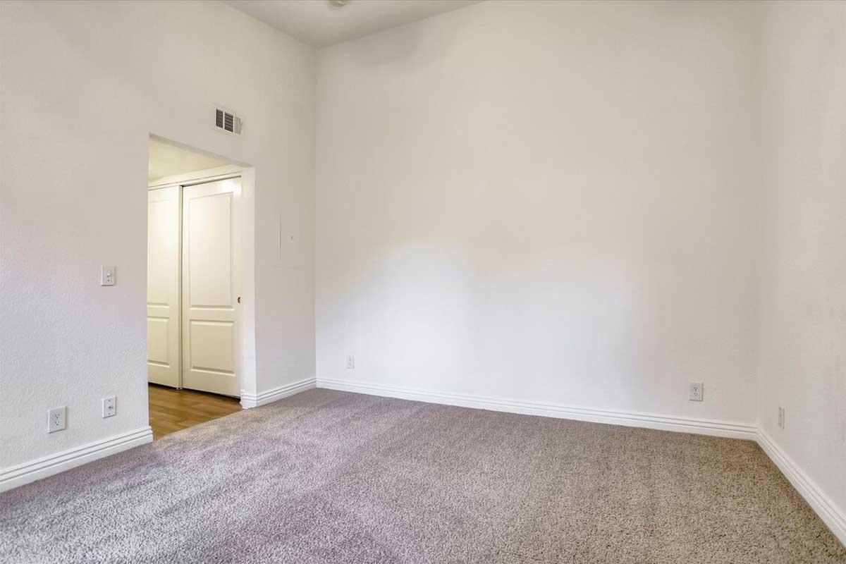 A spacious empty room with light-colored walls and plush carpet. A doorway on the left leads to another room with white panel doors, and the room features a high ceiling and ample natural light. The overall ambiance is bright and inviting, ideal for personalizing or decorating.