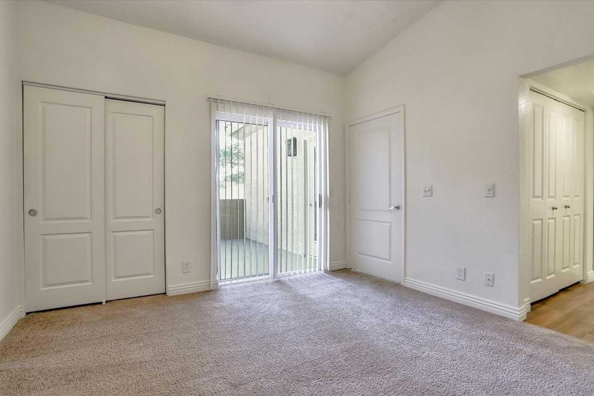 Interior view of a room featuring light-colored walls and carpeted flooring. There are two closed closet doors on the left, and large sliding glass doors leading to a balcony on the right. The room is well-lit with natural light and has a simple, modern design. A door on the right leads to another area.