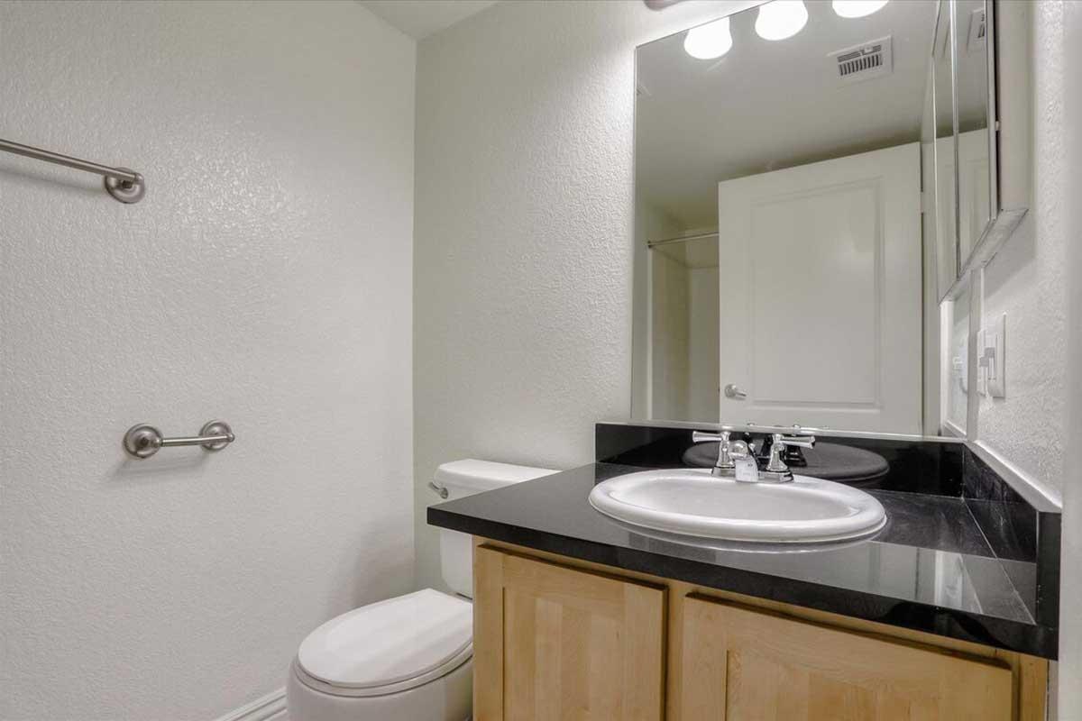 A clean, minimalistic bathroom featuring a black countertop with a white sink, a mirror above it, a white toilet, and silver grab bars. The walls are painted light, and there is a towel rack nearby, contributing to a bright and modern aesthetic.