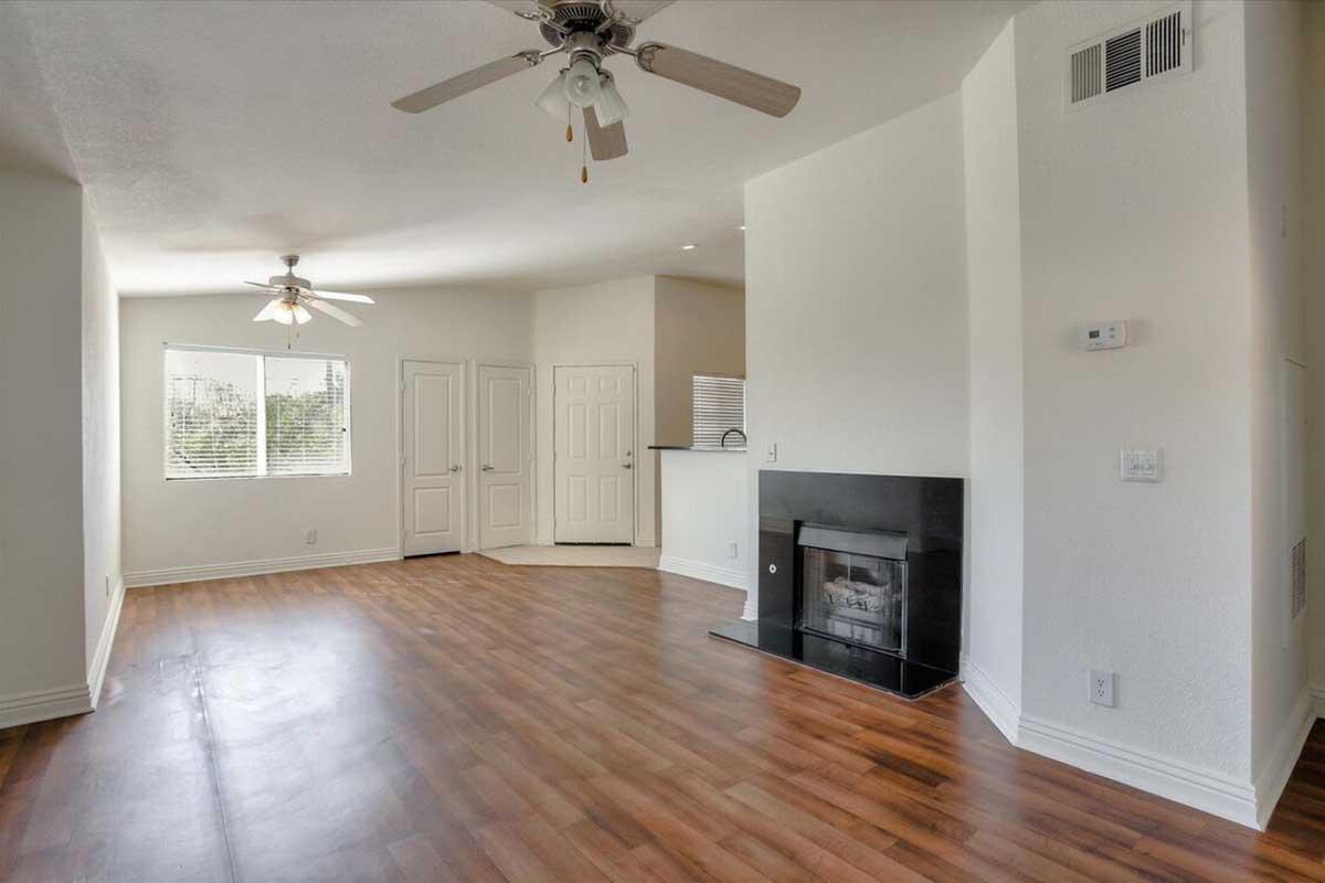 Spacious living room with hardwood floors, featuring a ceiling fan and a cozy fireplace. Two windows provide natural light, and a doorway leads to a closed room. The adjoining area suggests a kitchen with a bar counter viewable from the living space. Walls are painted in light neutral tones.