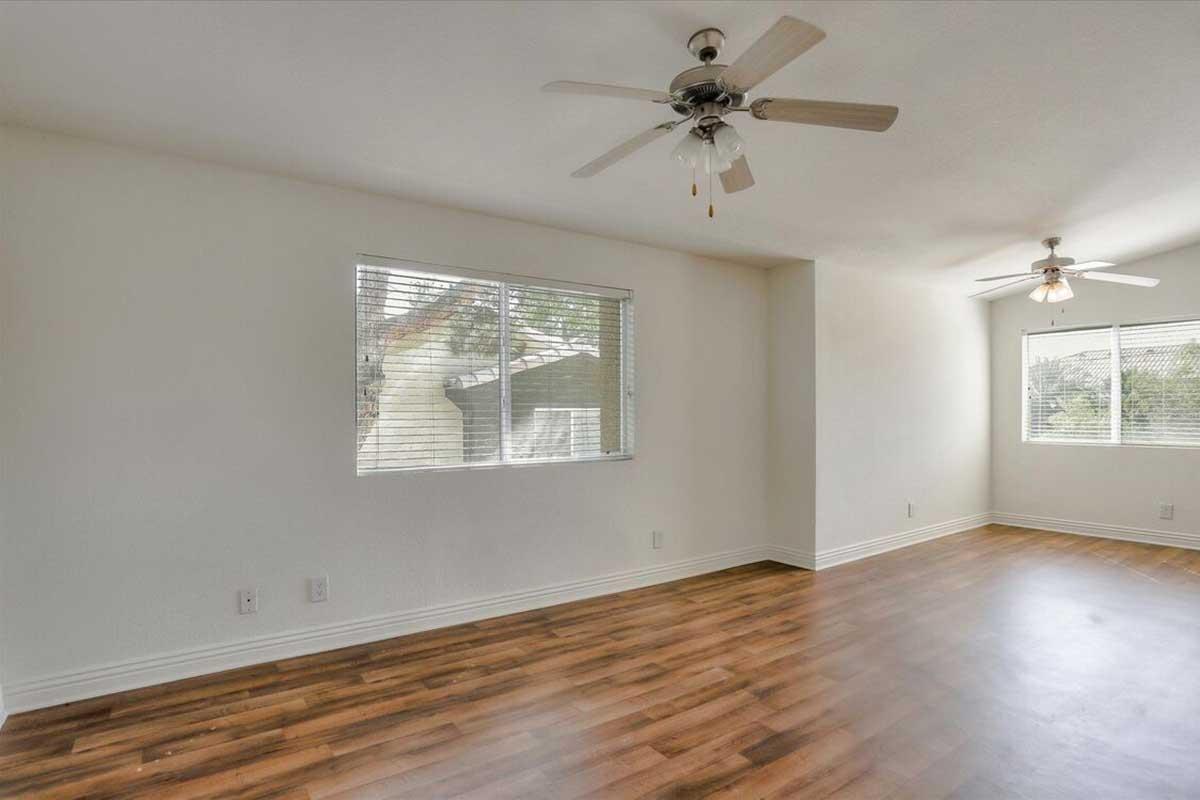 A bright and airy room with light-colored walls and wooden flooring. There are two ceiling fans and two windows allowing natural light to enter. The space appears spacious and uncluttered, suitable for various uses.