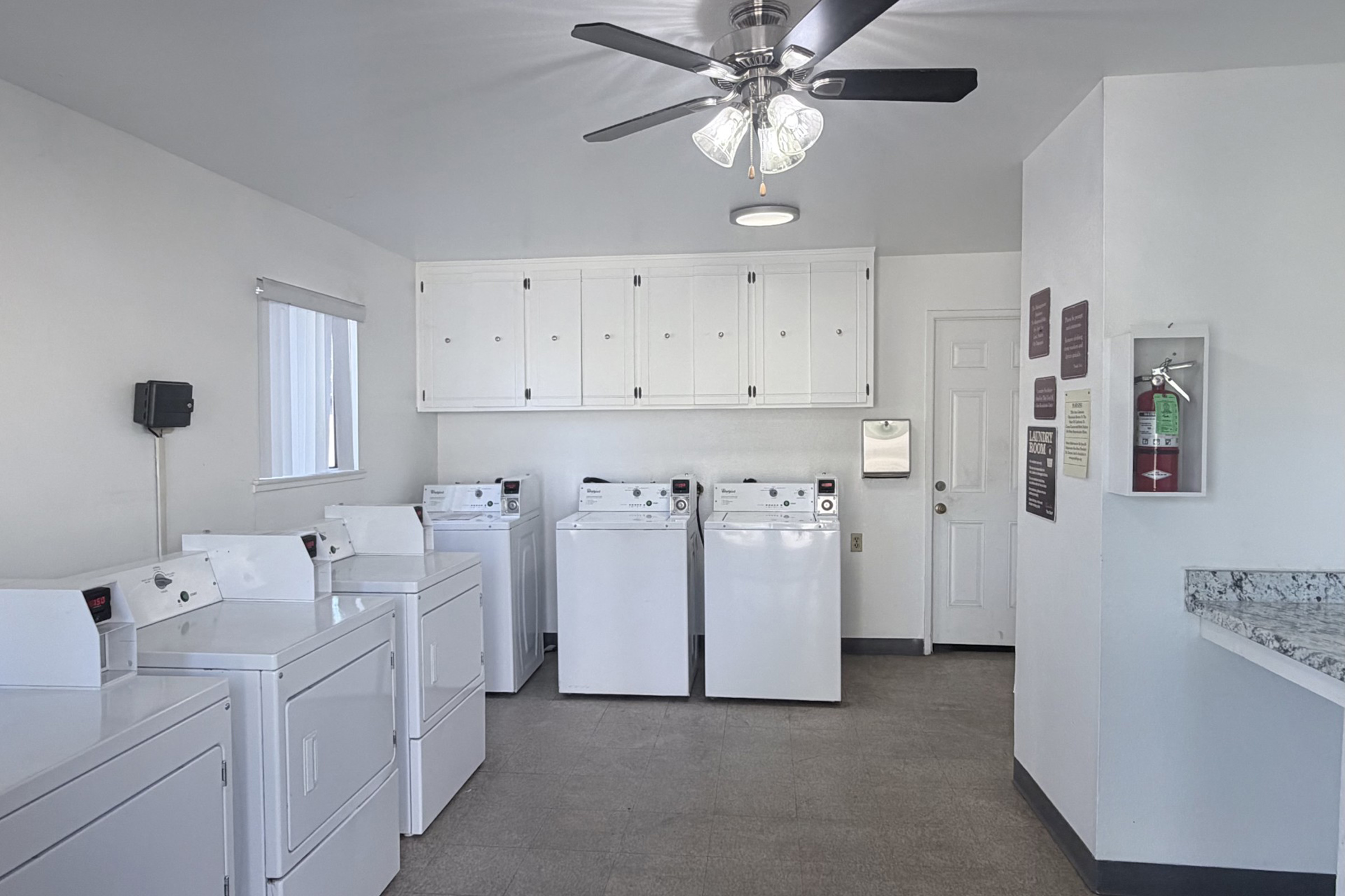 A clean laundry room featuring several white washing machines and dryers lined up against a wall. There is a ceiling fan, a window with blinds, and cabinets above the machines. A fire extinguisher is mounted on the wall, and there are informational signs posted by the entrance. The floor is tiled, and the walls are painted white.