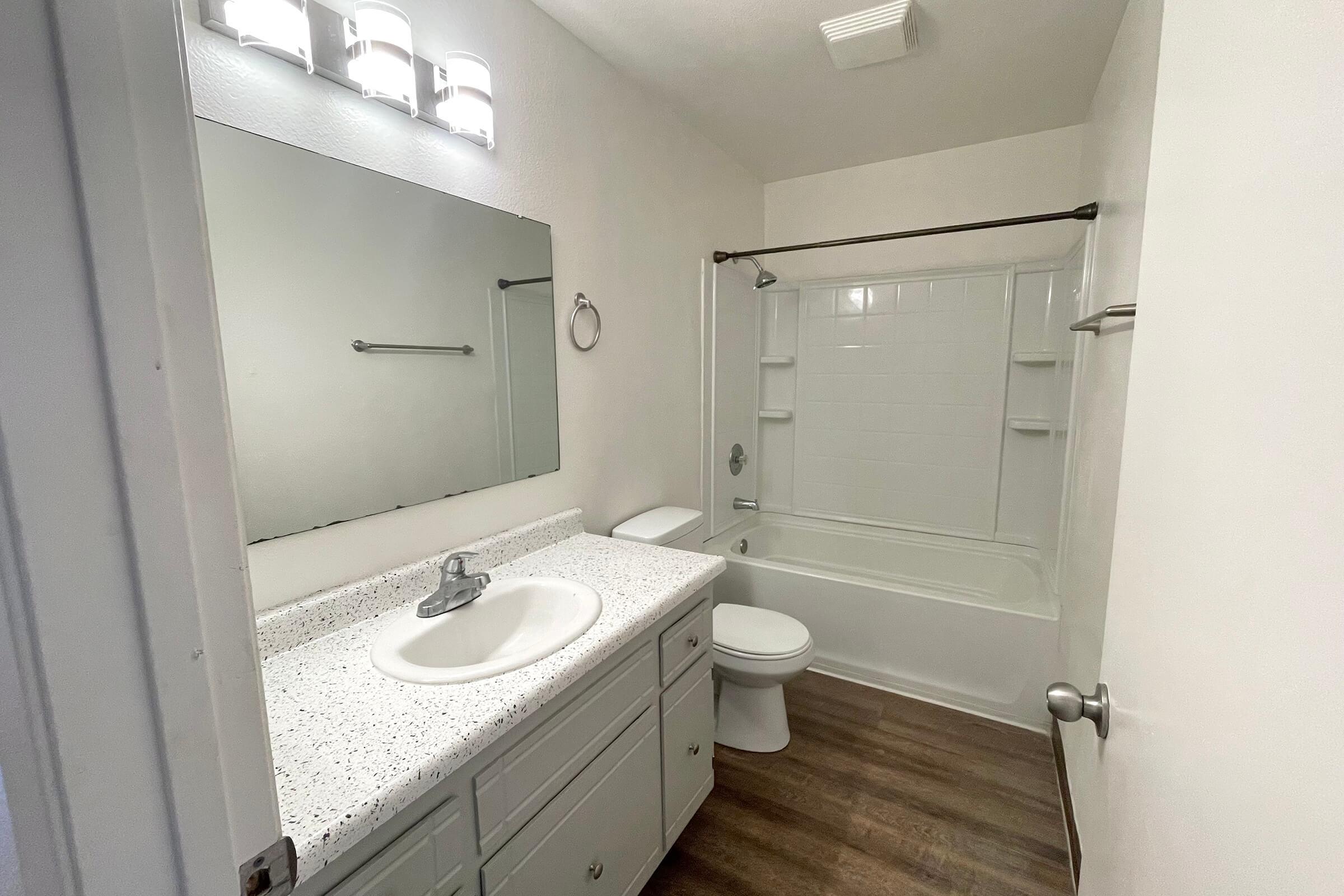 A clean, modern bathroom featuring a white bathtub with a shower, a large mirror above a white sink with a speckled countertop, and a toilet. The walls are painted in a light color, and there is a light fixture with three bulbs above the mirror. The floor has a wood-like appearance.