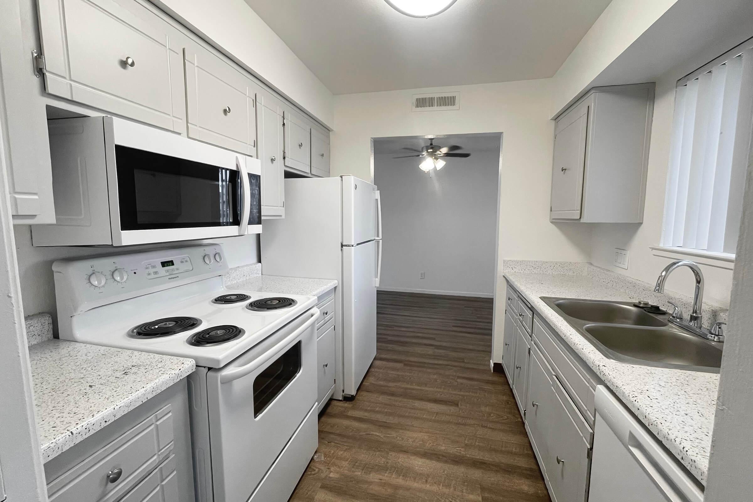Modern kitchen with white cabinetry and granite countertops. It features an oven, a microwave, and a refrigerator. There are dual sinks and a large window letting in natural light. The kitchen connects to a living area with a fan and has wooden flooring.