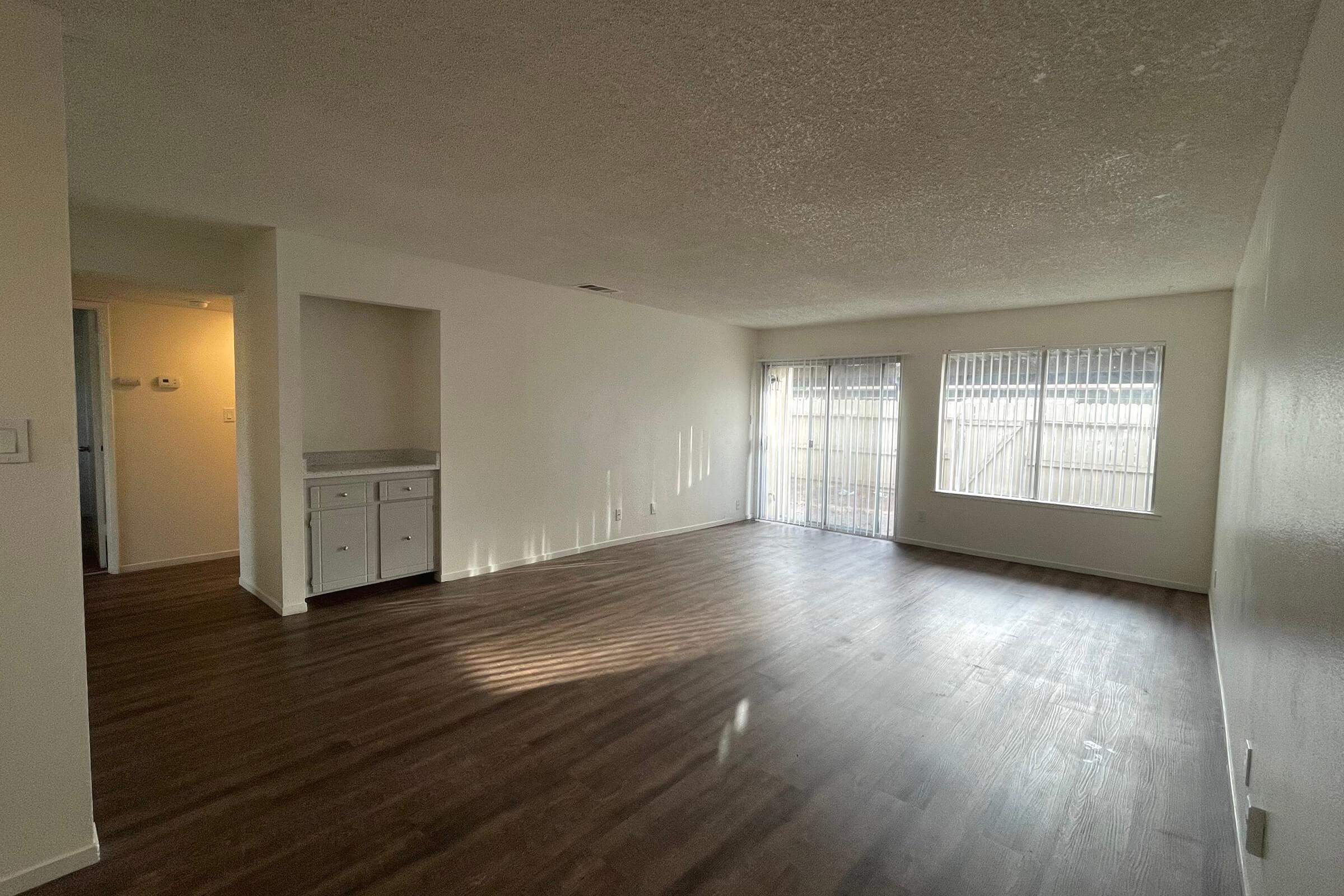 A spacious, empty living room with light-colored walls and a textured ceiling. It features hardwood floors and large windows with blinds, allowing natural light to enter the room. A small built-in cabinet is visible on the left wall, and there is an entrance to another room on the far left.