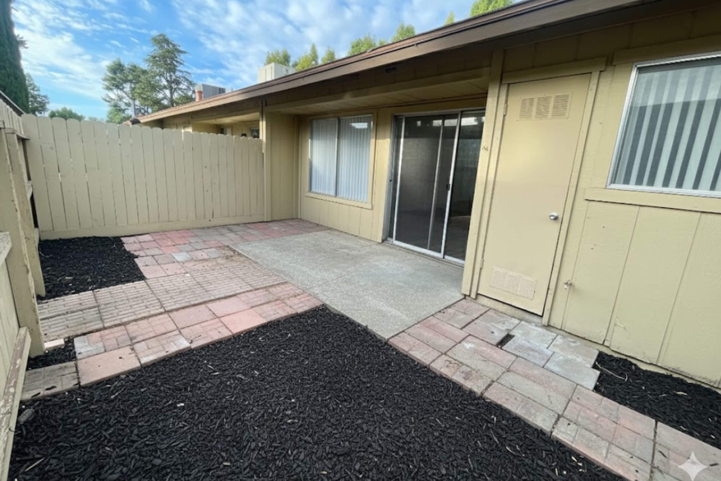 A small outdoor patio area with a concrete floor, bordered by paver stones and black mulch. There is a wooden fence in the background, and sliding glass doors leading into a room. The sky is partly cloudy, and some trees are visible in the distance.