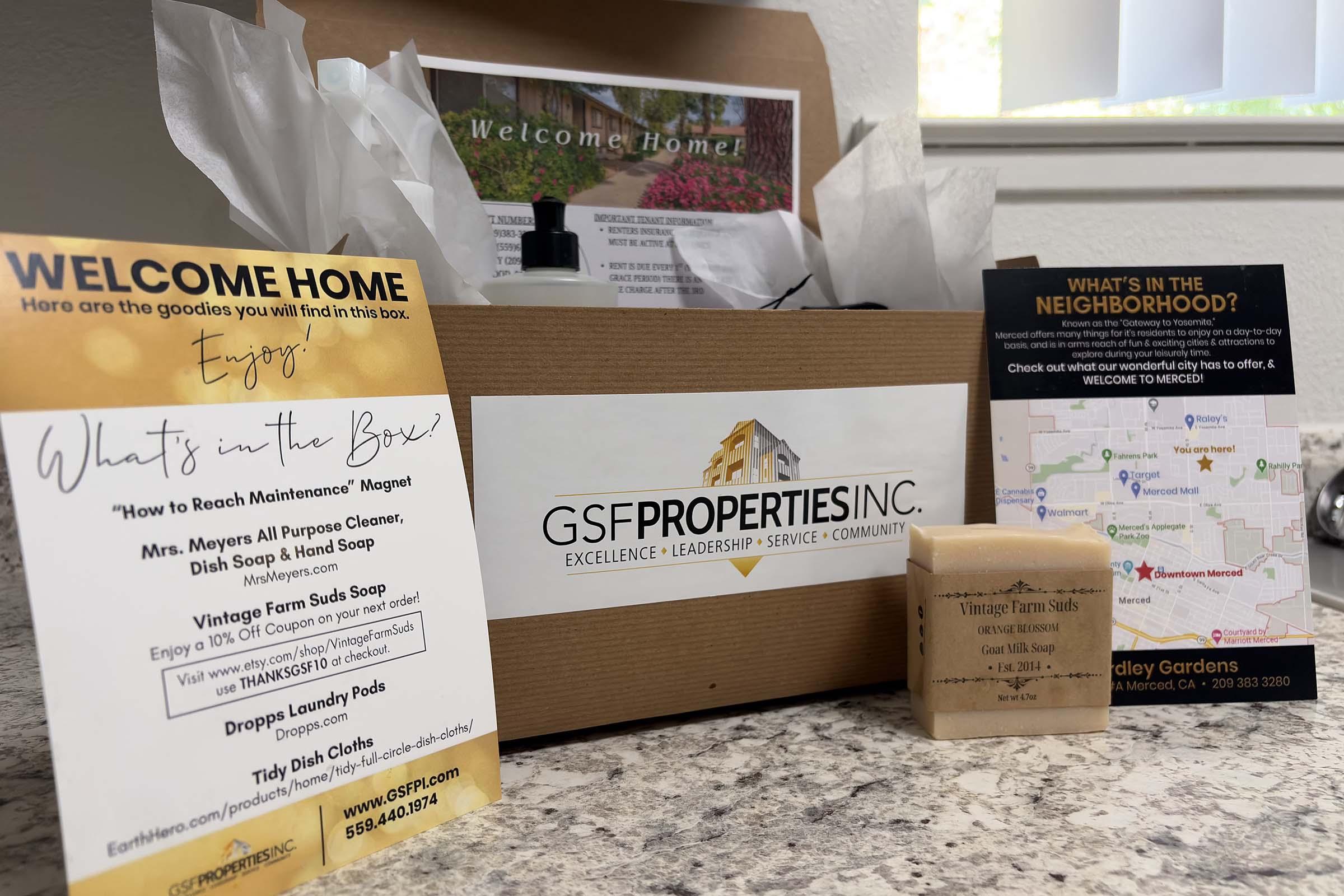 A welcome home gift box from GSF Properties Inc. on a countertop, featuring a welcome note, promotional materials, and a bar of soap. The box contains items such as a cleaner and hand soap, with additional flyers detailing local services and amenities. Decor includes crumpled tissue paper for presentation.
