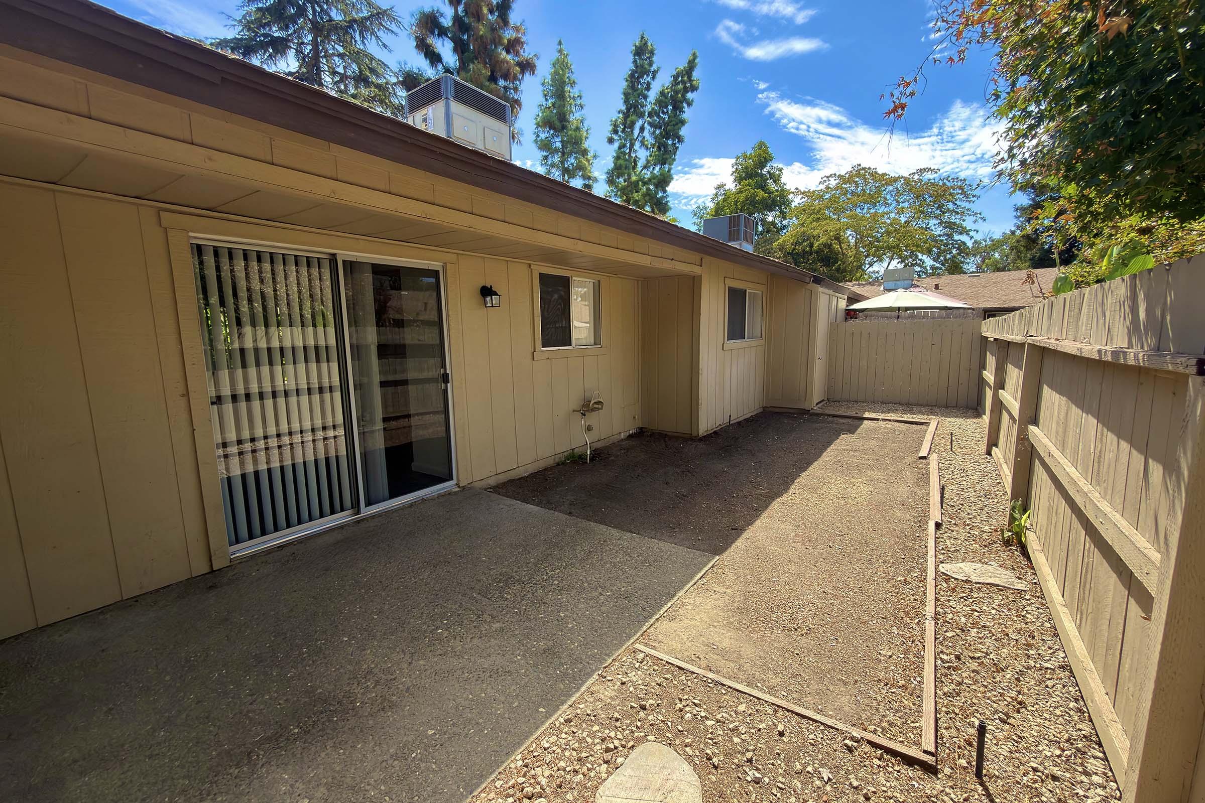 A view of the side yard of a single-story house featuring beige siding, a sliding glass door with vertical blinds, and a concrete pathway. There is a small plant next to the house, and the area is surrounded by a wooden fence and trees in the background under a clear blue sky.