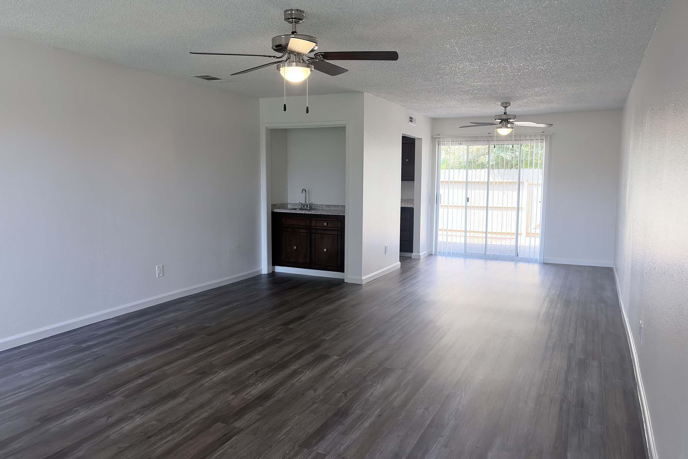 Spacious interior of a modern living room featuring light-colored walls and dark laminate flooring. Two ceiling fans provide airflow, and a small kitchenette area is visible to the left. The room has large windows that allow natural light to fill the space, leading to an outdoor area with a patio.