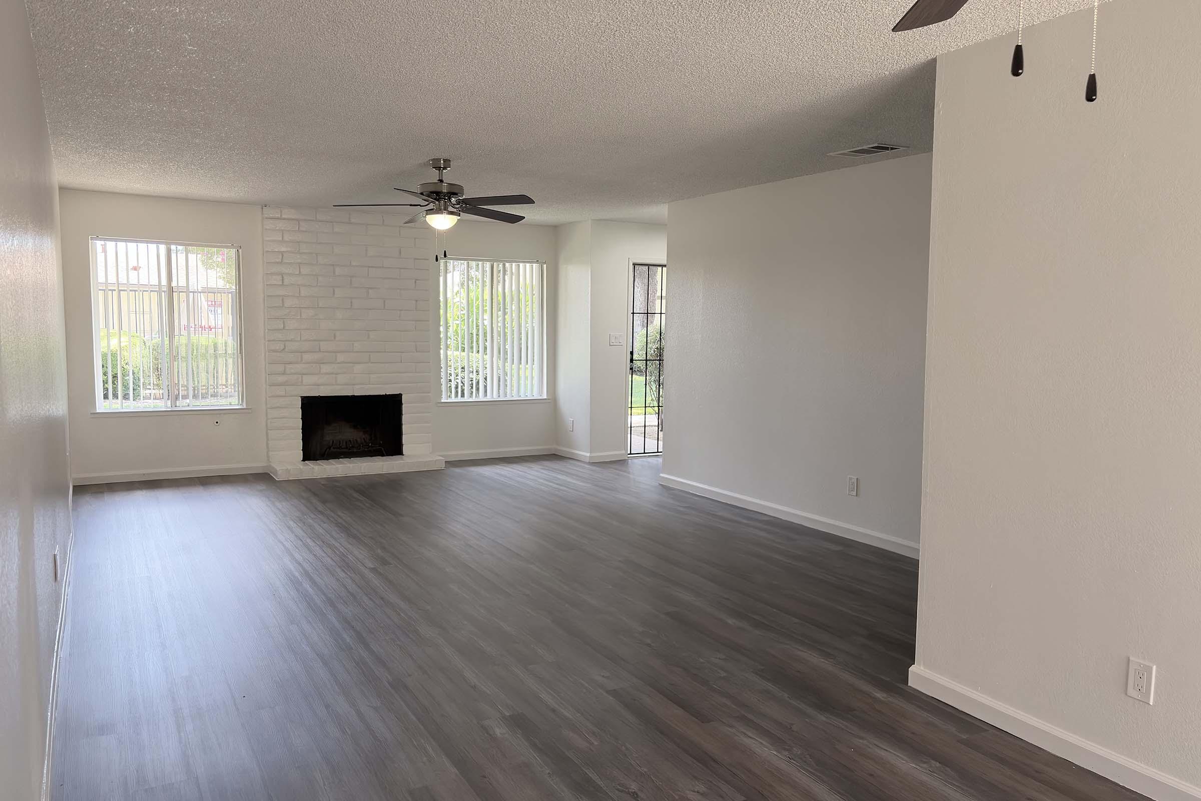 A spacious, well-lit living room with a ceiling fan and large windows. The flooring is a light-colored wood, and there's a white brick fireplace on one wall. The room features neutral paint and an open layout leading to another room visible in the background.