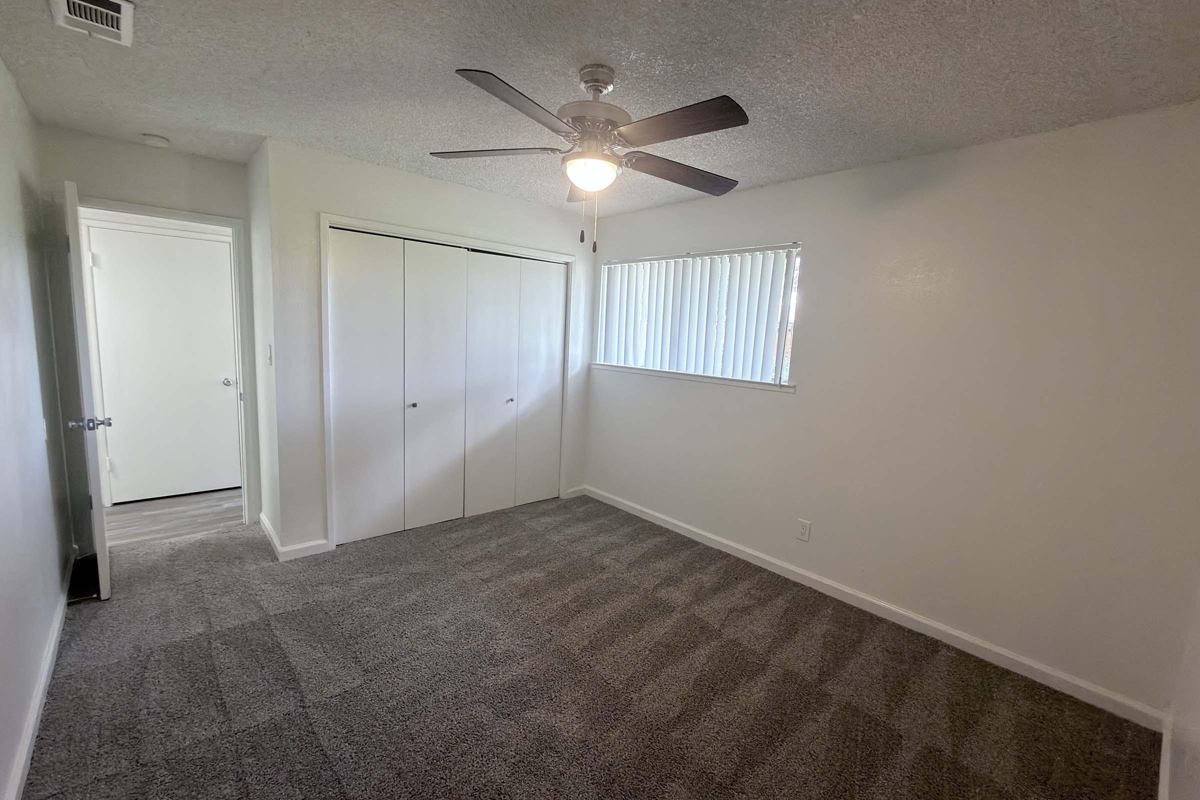 A well-lit room with light-colored walls and carpet. Flanked by a closet with sliding doors on one side, a window with blinds allows natural light. A ceiling fan is centered, and the doorway leads to a hallway. The space is clean and empty, suitable for various用途.